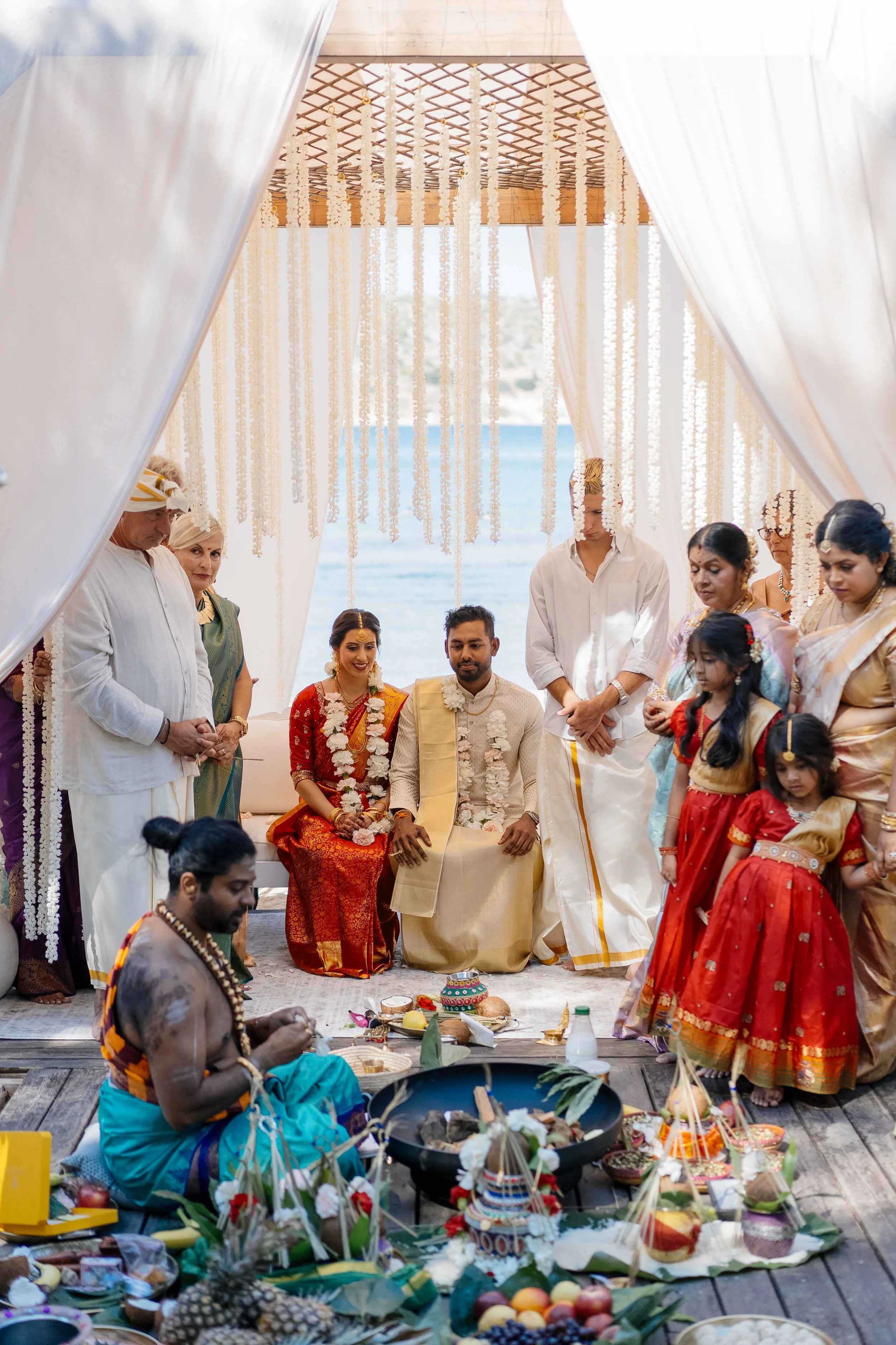 Hindu wedding ceremony with a priest performing rituals, a bride and groom seated in traditional attire, and family members and children dressed in colorful garments, under a decorated canopy overlooking the water.