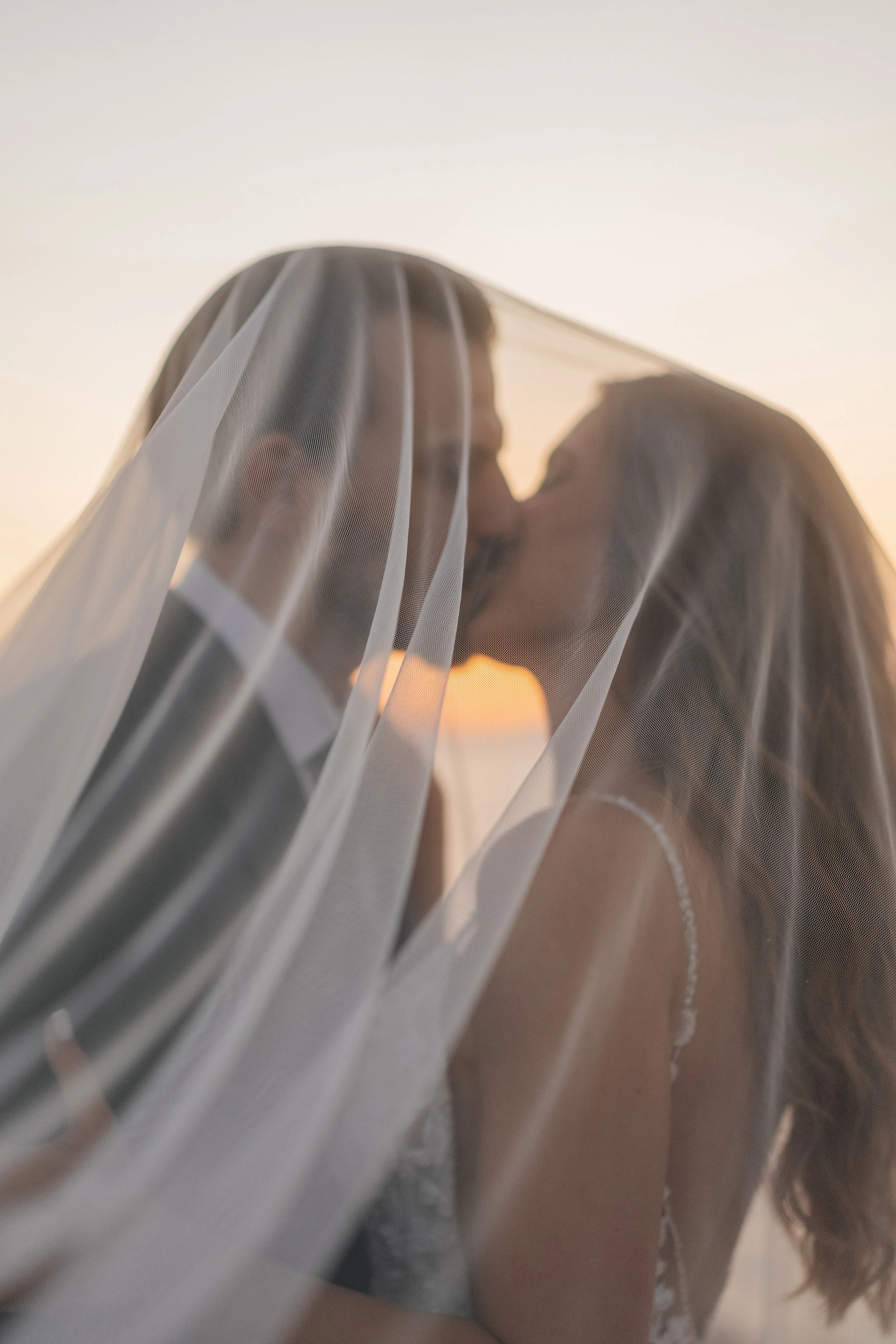 Two women sharing a kiss behind a sheer white curtain during sunset