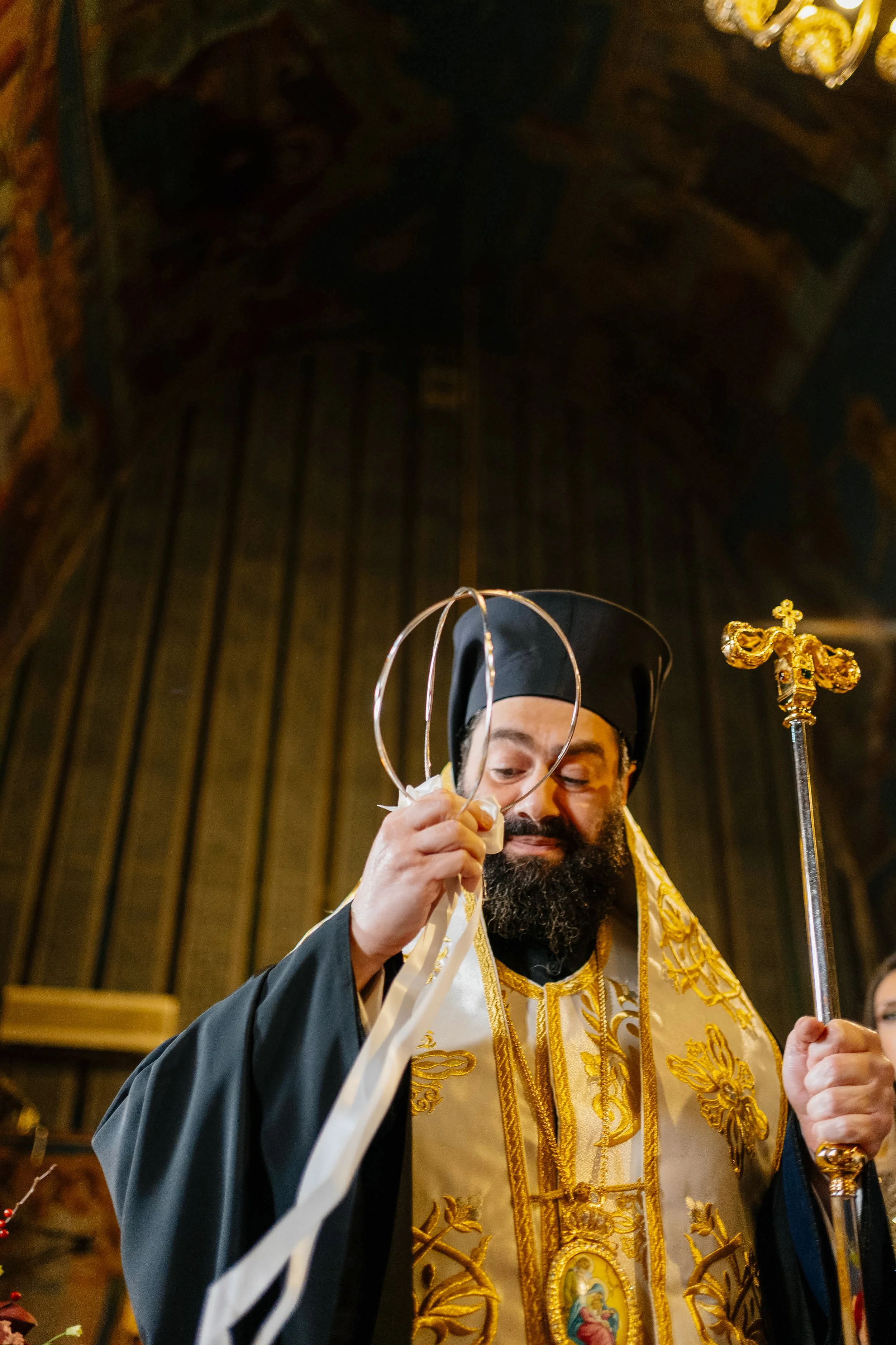 A man wearing religious robes and a mitre, holding a staff, standing in a church setting.