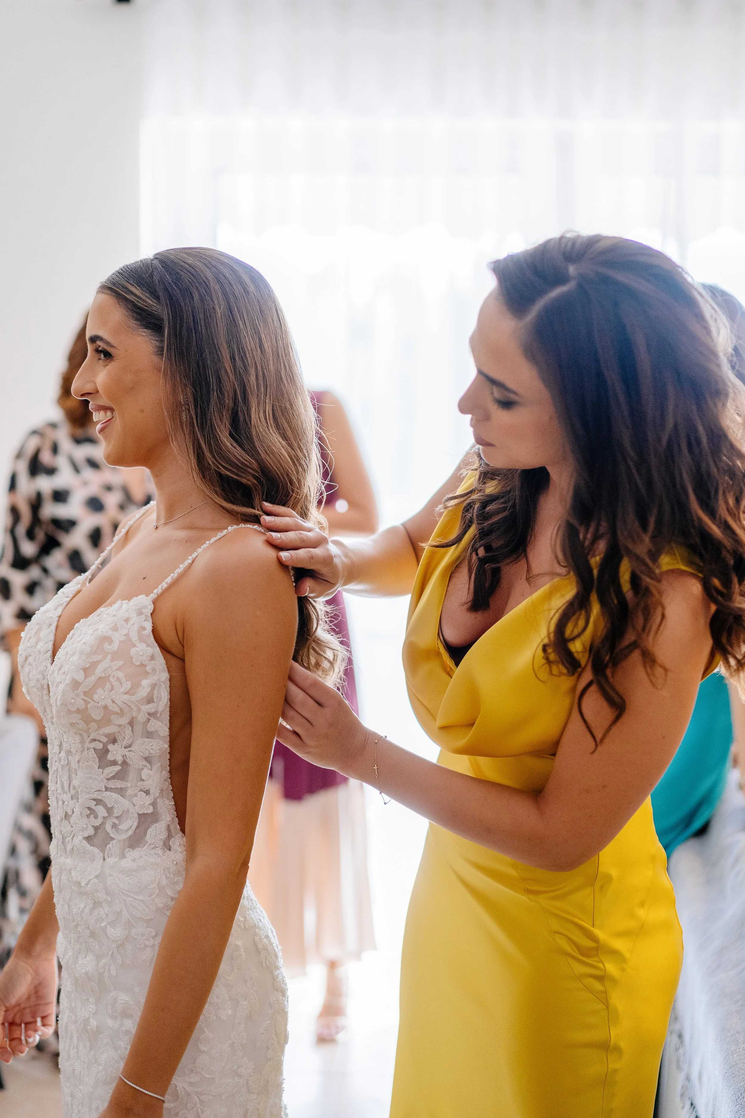 A woman in a white lace wedding dress smiling as she is helped into her dress by another woman in a yellow dress, with other women in colorful dresses in the background, in a bright room with white curtains.
