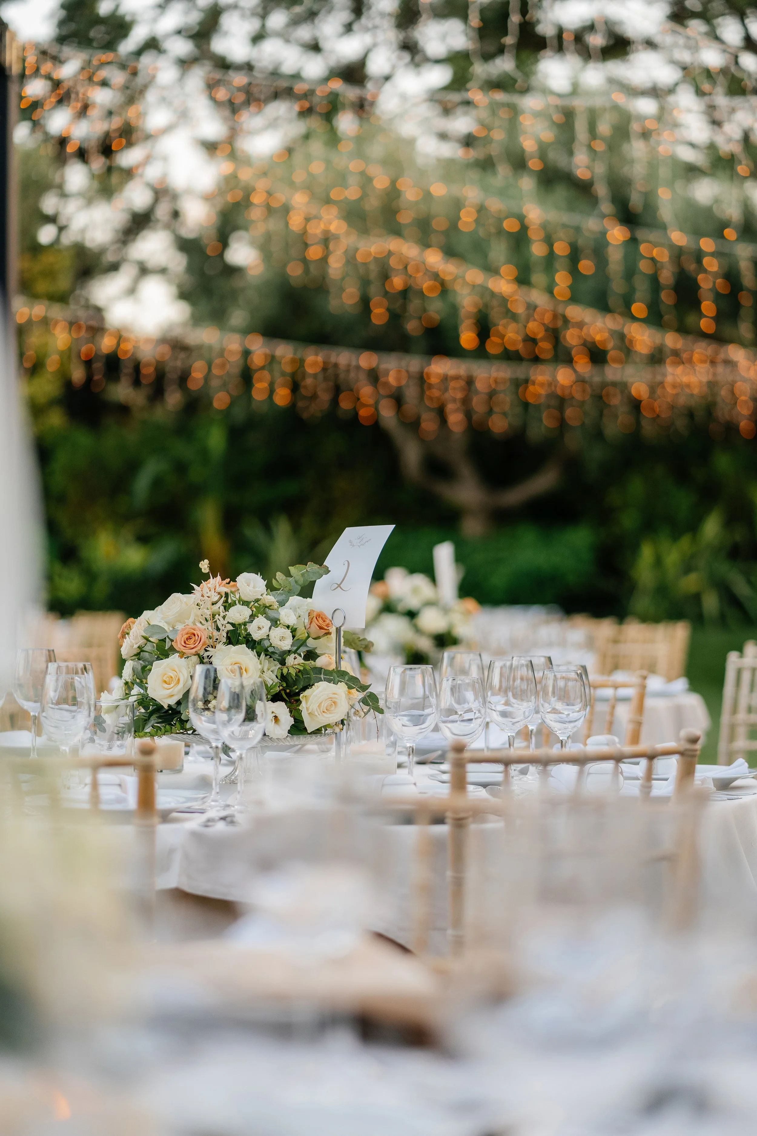 A wedding reception table decorated with a flower arrangement of white and peach roses, surrounded by multiple empty wine glasses, set outdoors with a blurred background of string lights and trees.