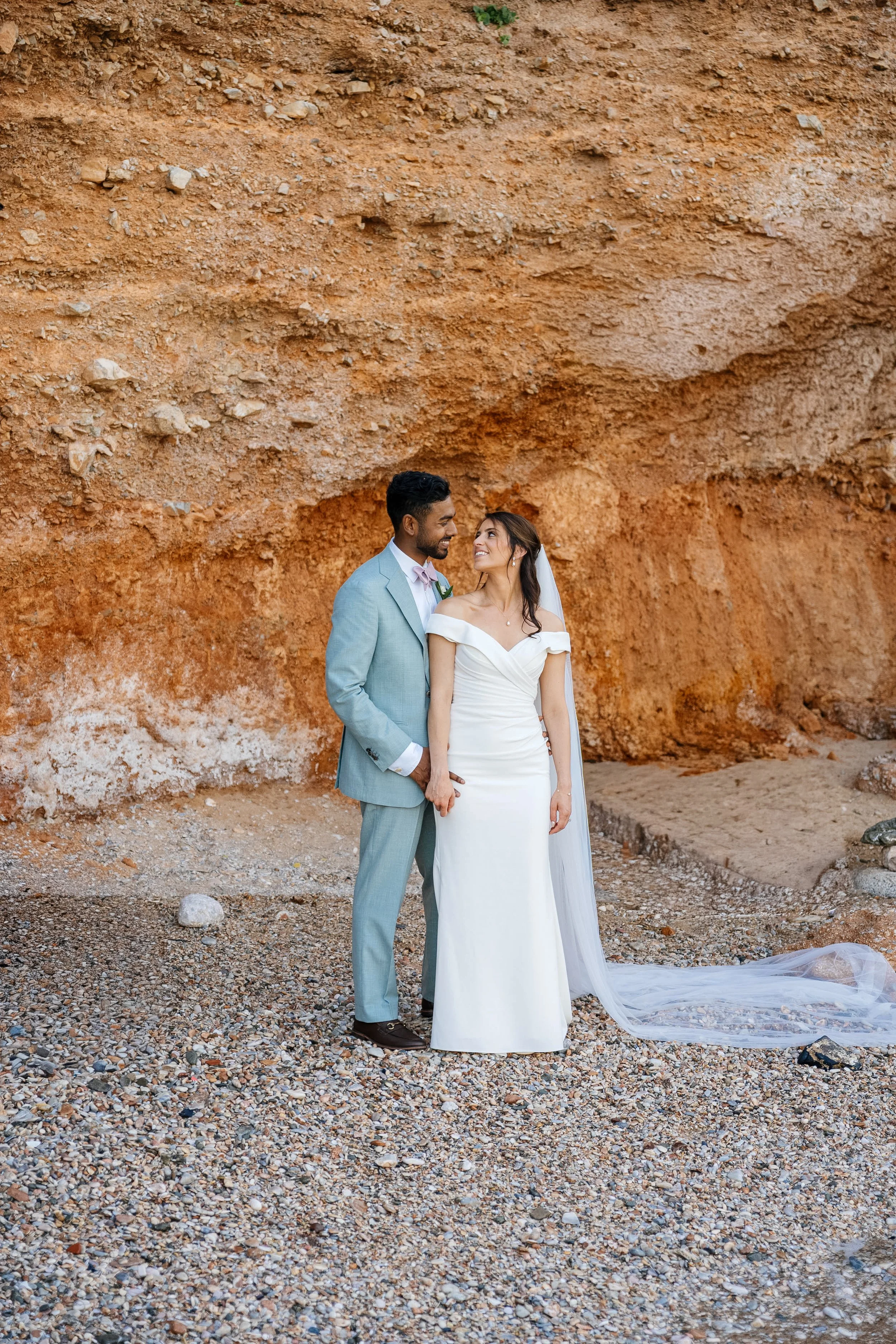 A bride and groom standing close together on a rocky beach with a large red rock formation in the background. The groom is wearing a light blue suit with a pink tie, and the bride is in a white off-the-shoulder wedding gown with a veil. They are look