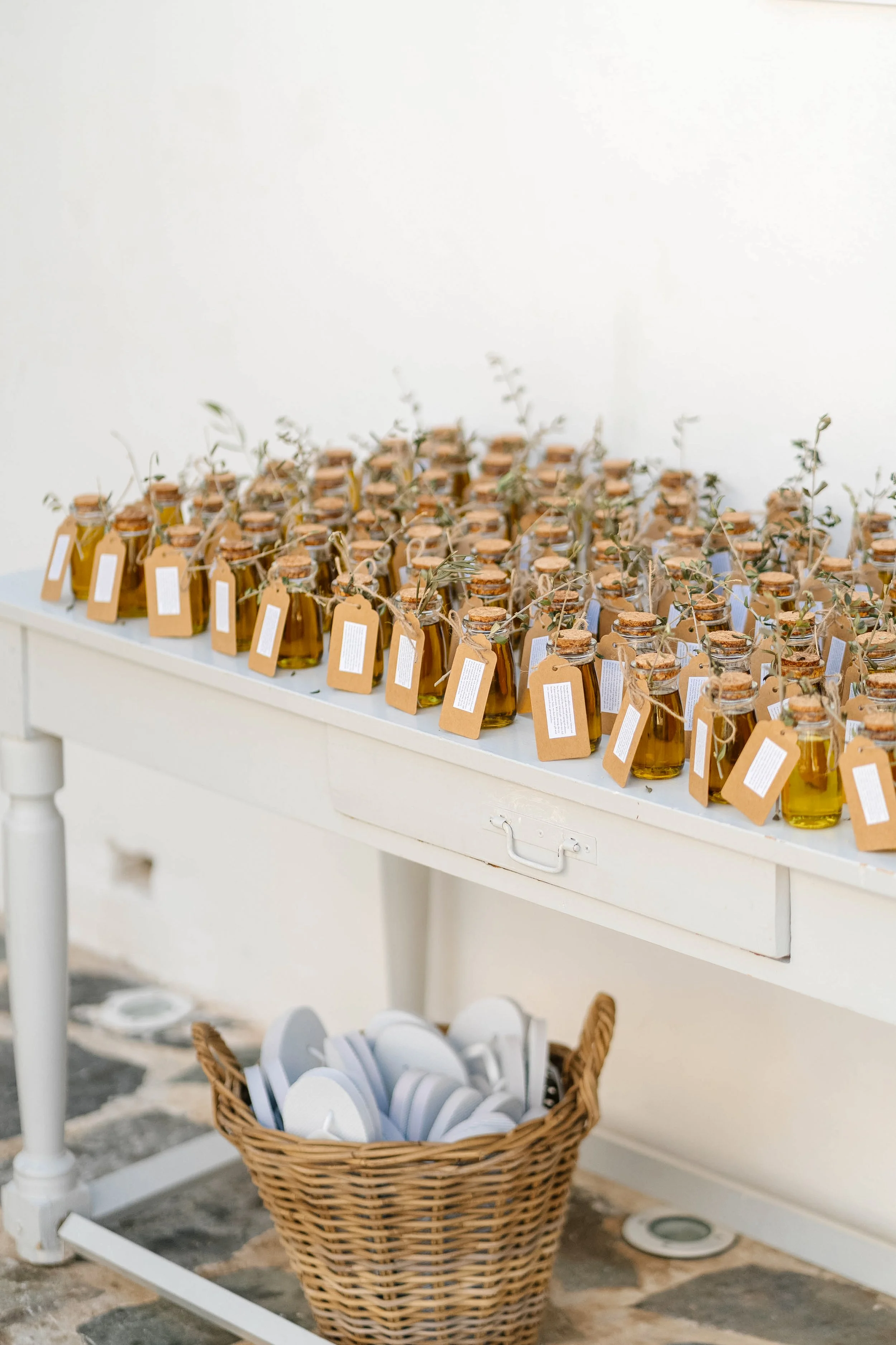 A white table with small glass bottles filled with yellow liquid, each sealed with a cork and tagged with a brown paper label, decorated with sprigs of greenery. Below the table, a wicker basket holds white earbuds, and a white wall in the background