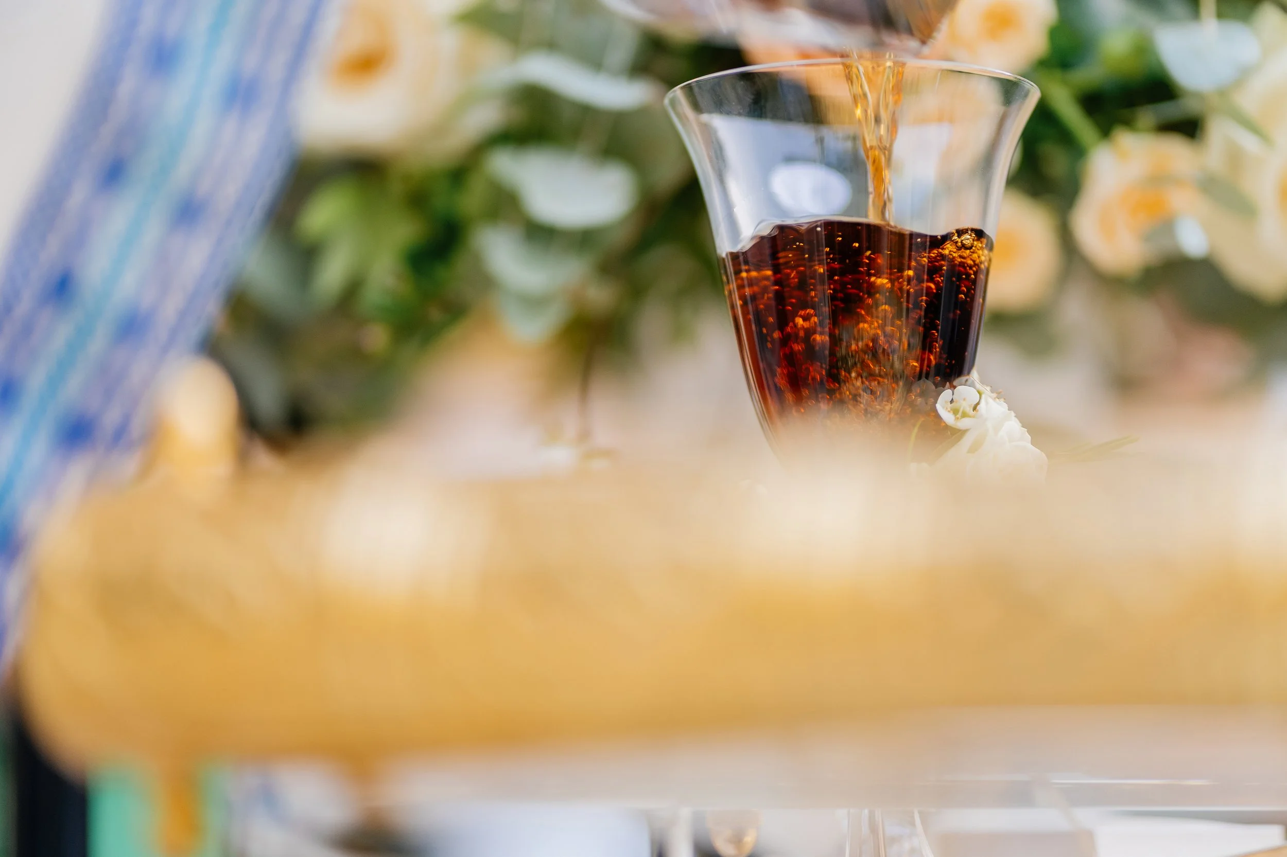 A glass of dark soda with bubbles being poured into it from a bottle, with green and white blurred flowers in the background.