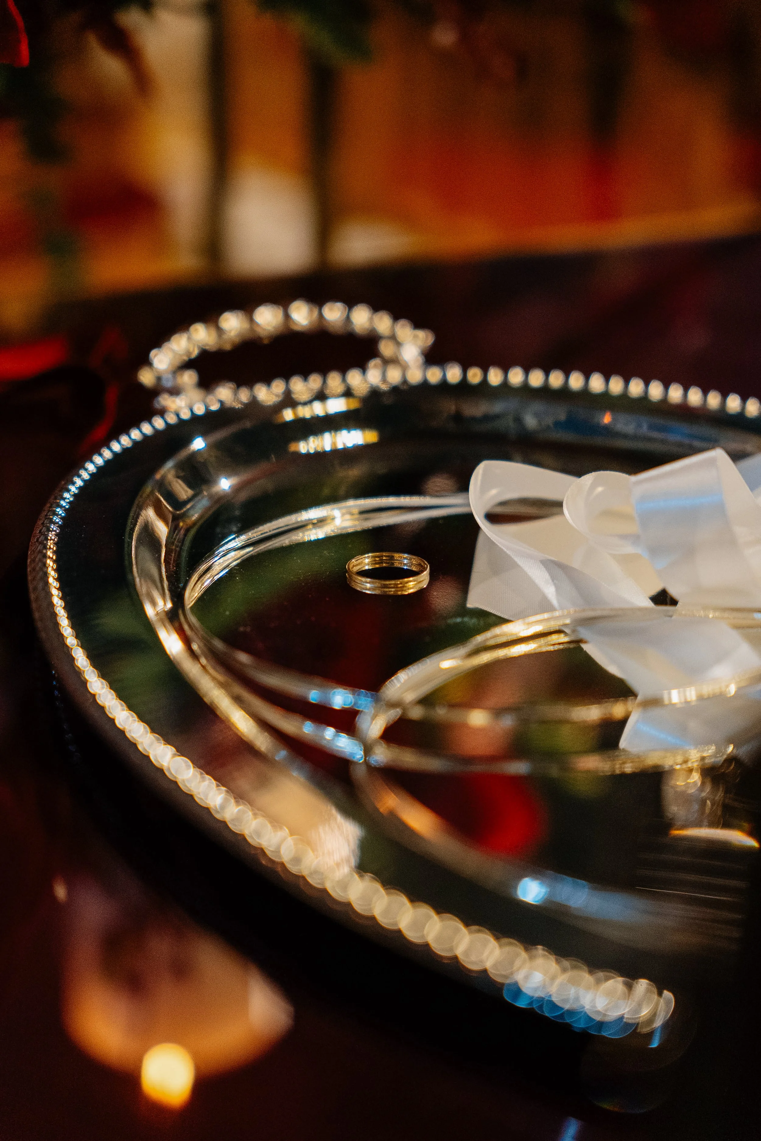 A blurred close-up image of jewelry, possibly a ring, with a shiny metallic surface and small clear stones, along with a white ribbon or cloth, set against a dark background with warm-tone reflections.