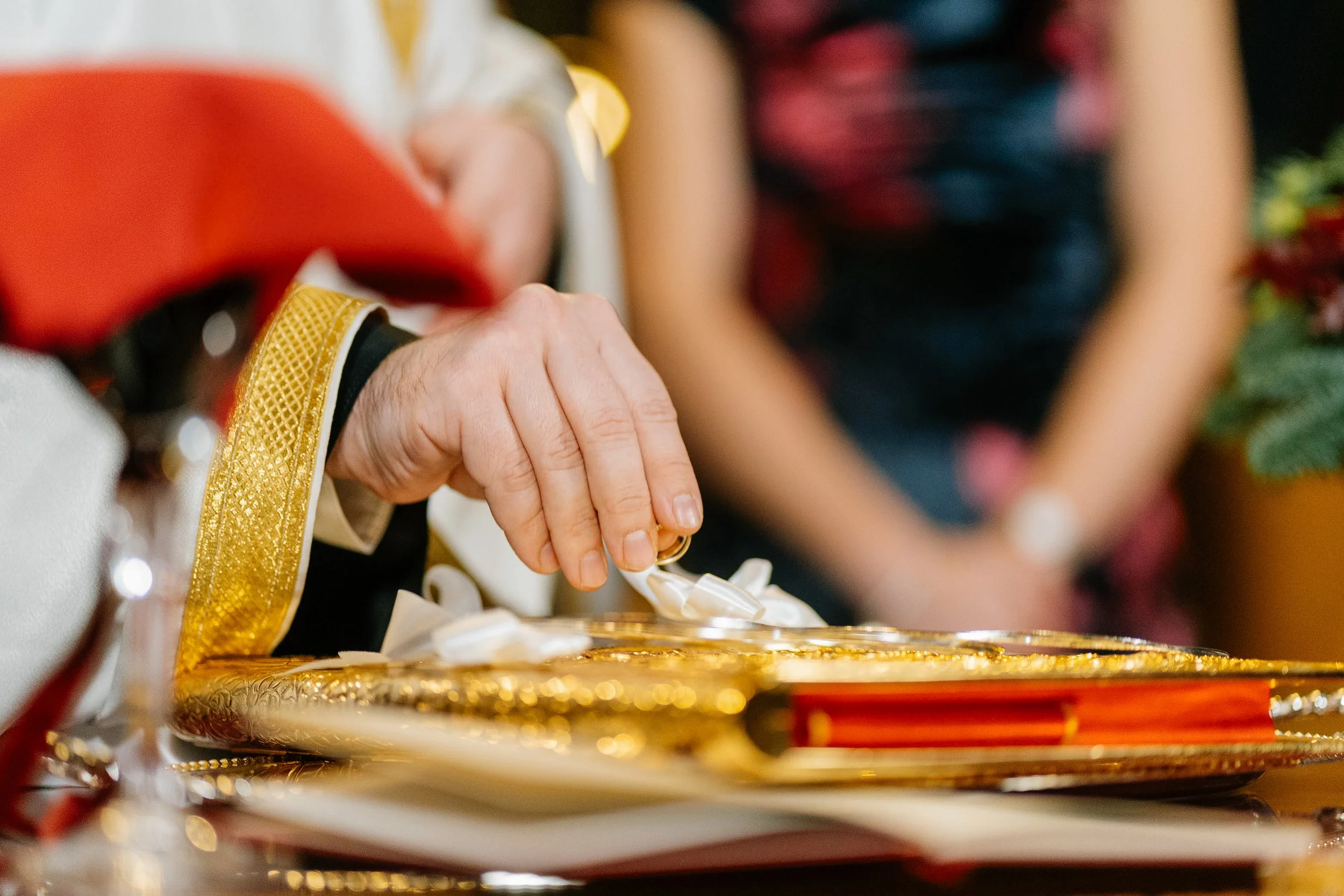 Close-up of a hand with a gold ring, touching a golden tray with decorative edges, with blurred people in the background.