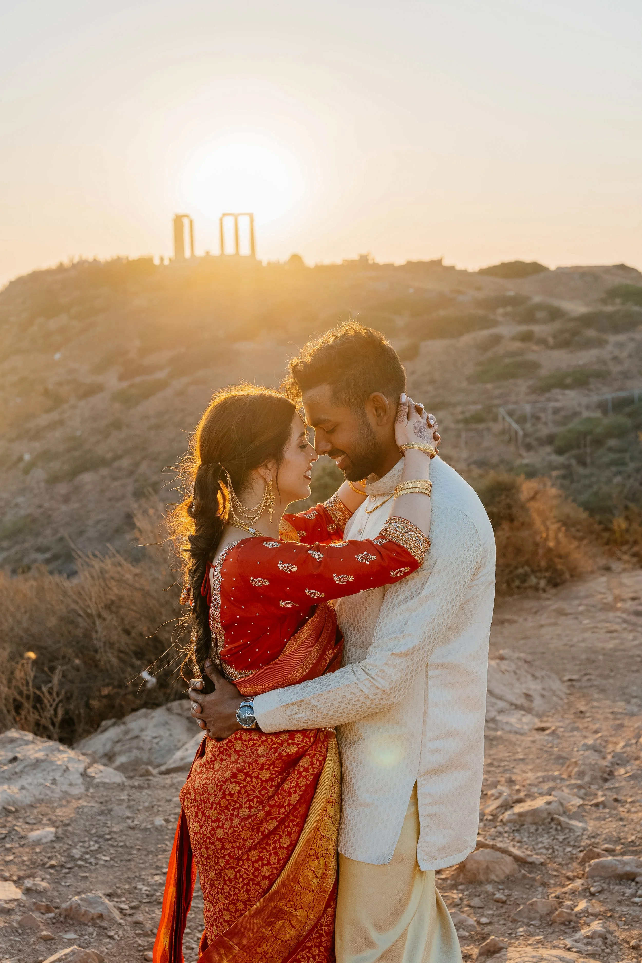 A couple dressed in traditional Indian attire sharing an intimate moment at sunset, standing on rocky terrain with hills in the background.