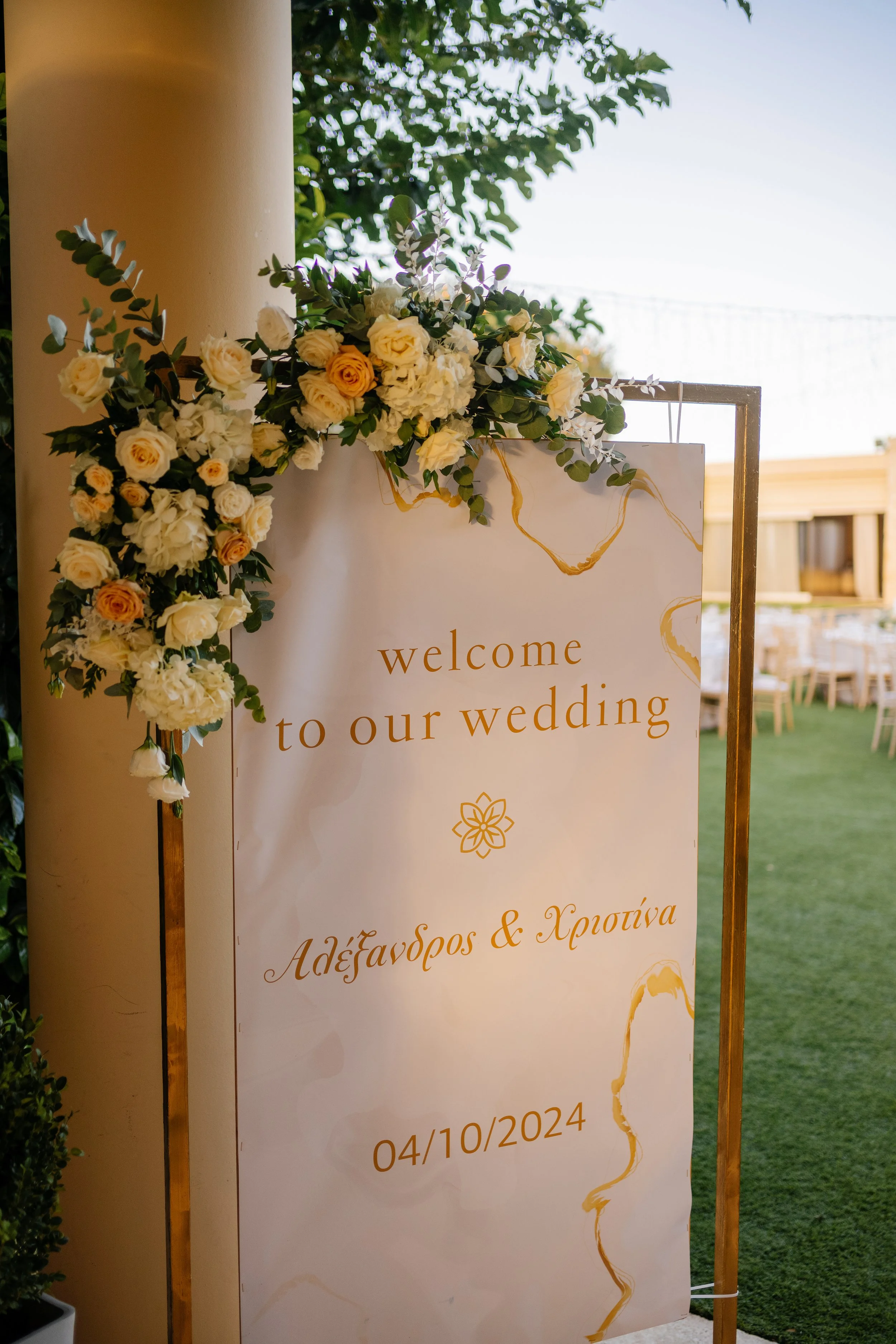 Wedding welcome sign decorated with white and cream flowers, greenery, and gold accents, displaying the date 04/10/2024, and written in both English and Greek.
