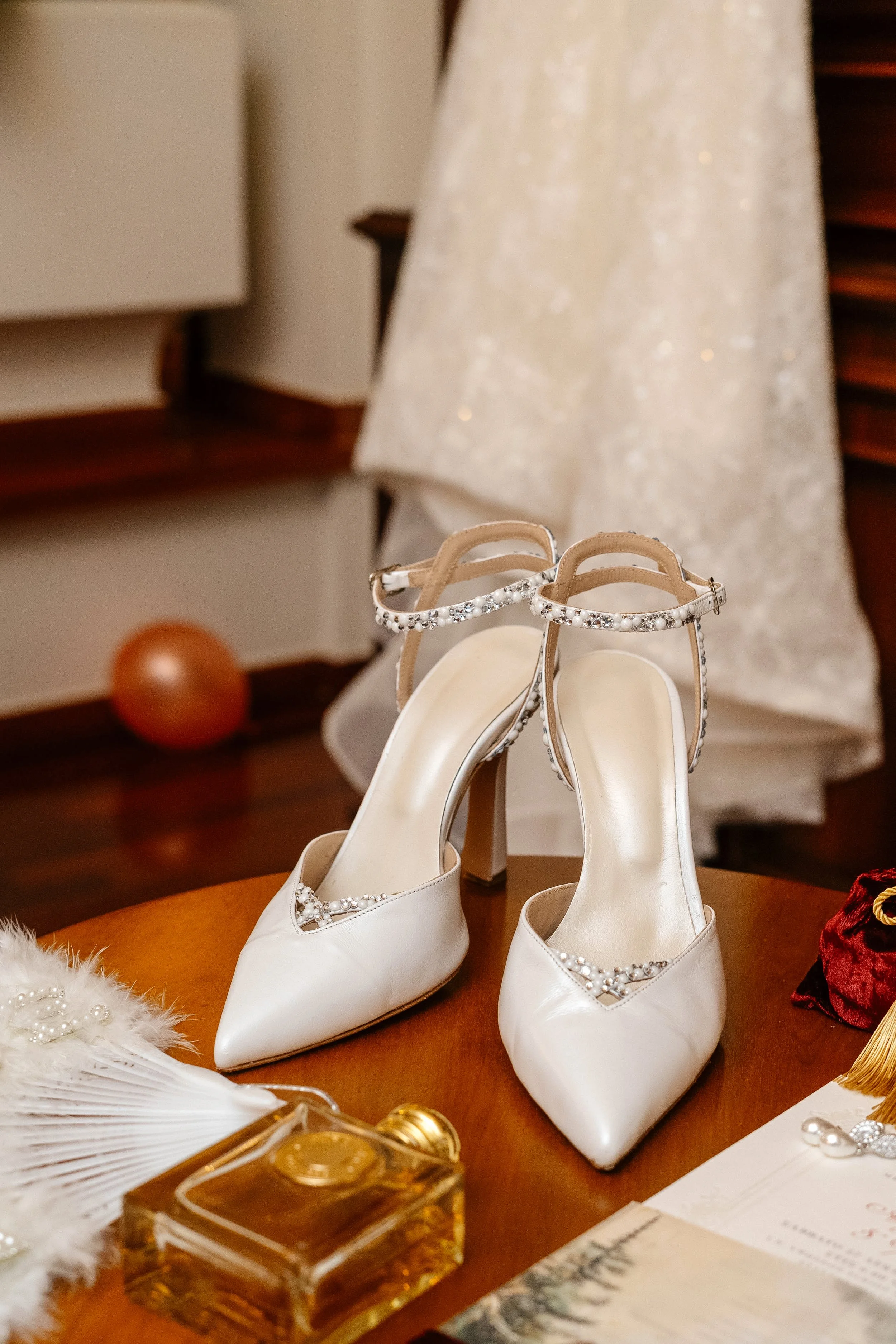 Elegant white bridal shoes with pearl embellishments on a wooden table, surrounded by vintage-style accessories.