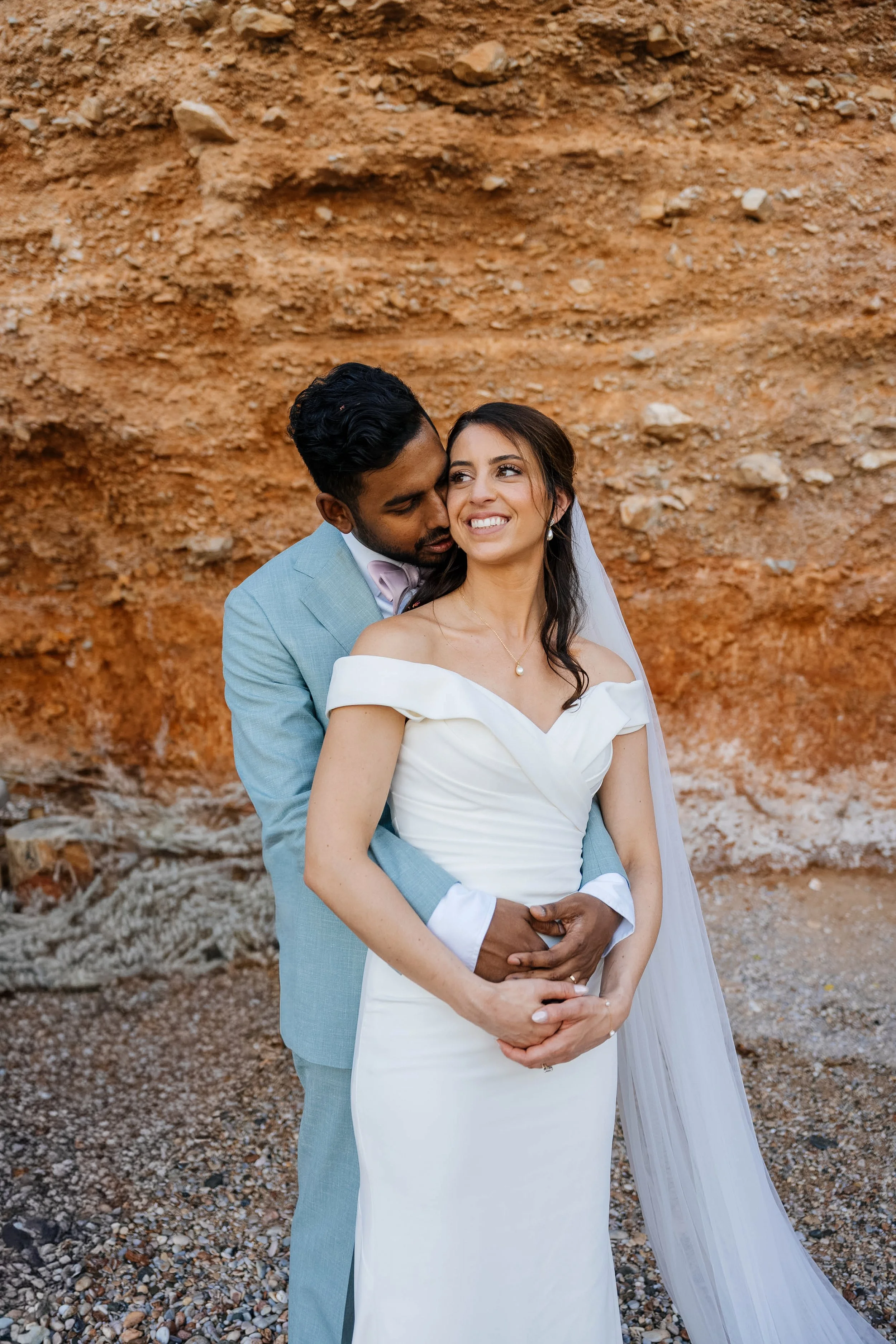 A couple dressed in wedding attire standing outdoors in front of a rocky, dirt background. The man is wearing a light blue suit and the woman is in a white off-the-shoulder wedding dress, smiling happily.