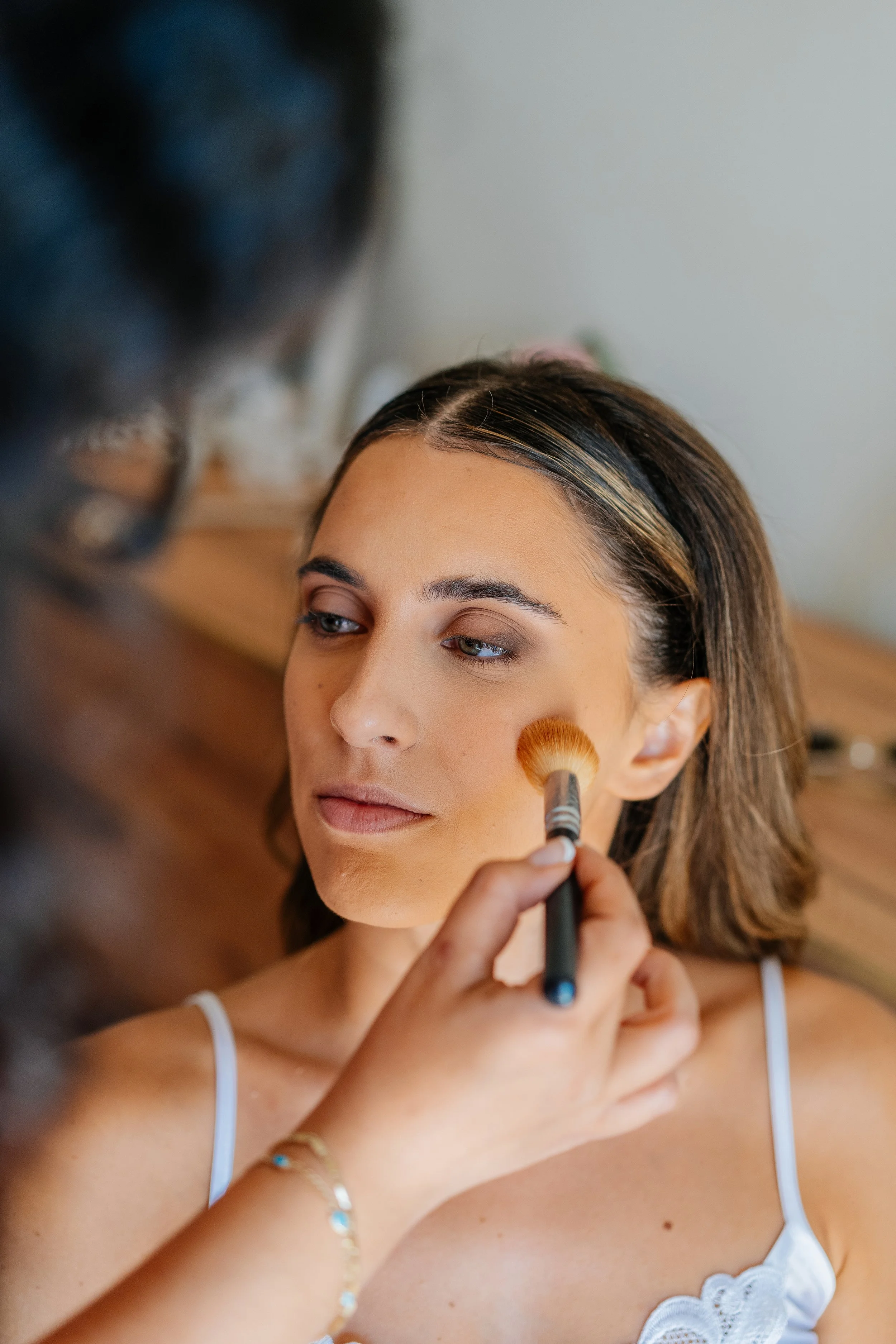 A woman with shoulder-length brown hair and light makeup is having her makeup done by a makeup artist using a fluffy brush on her cheek. The woman is wearing a white spaghetti strap top and a delicate bracelet with blue stones.