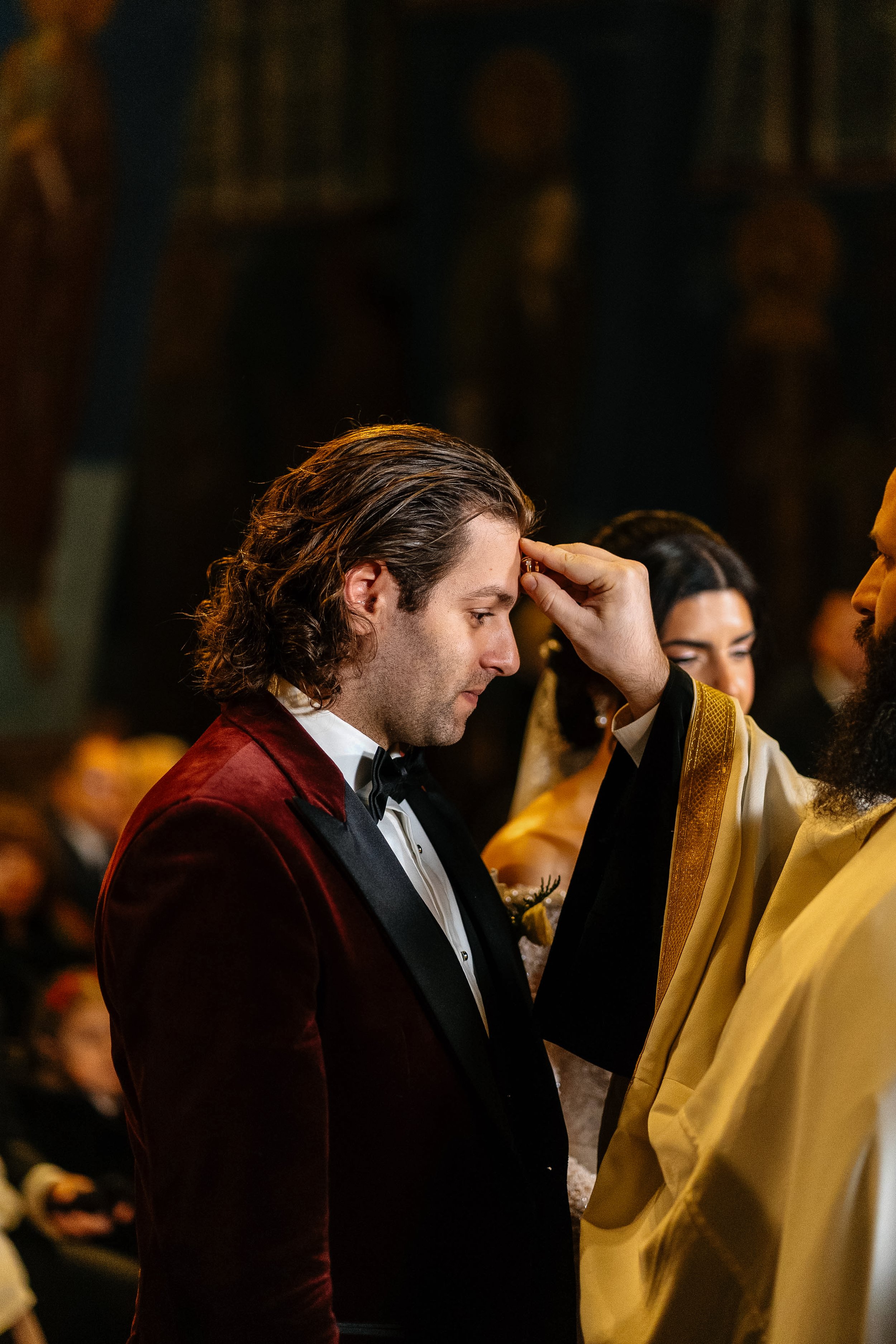 A man in a tuxedo and burgundy velvet jacket kneeling with a woman to be baptized, who is wearing ceremonial robes, in a church setting. The man is touching her forehead, possibly during a religious ceremony.