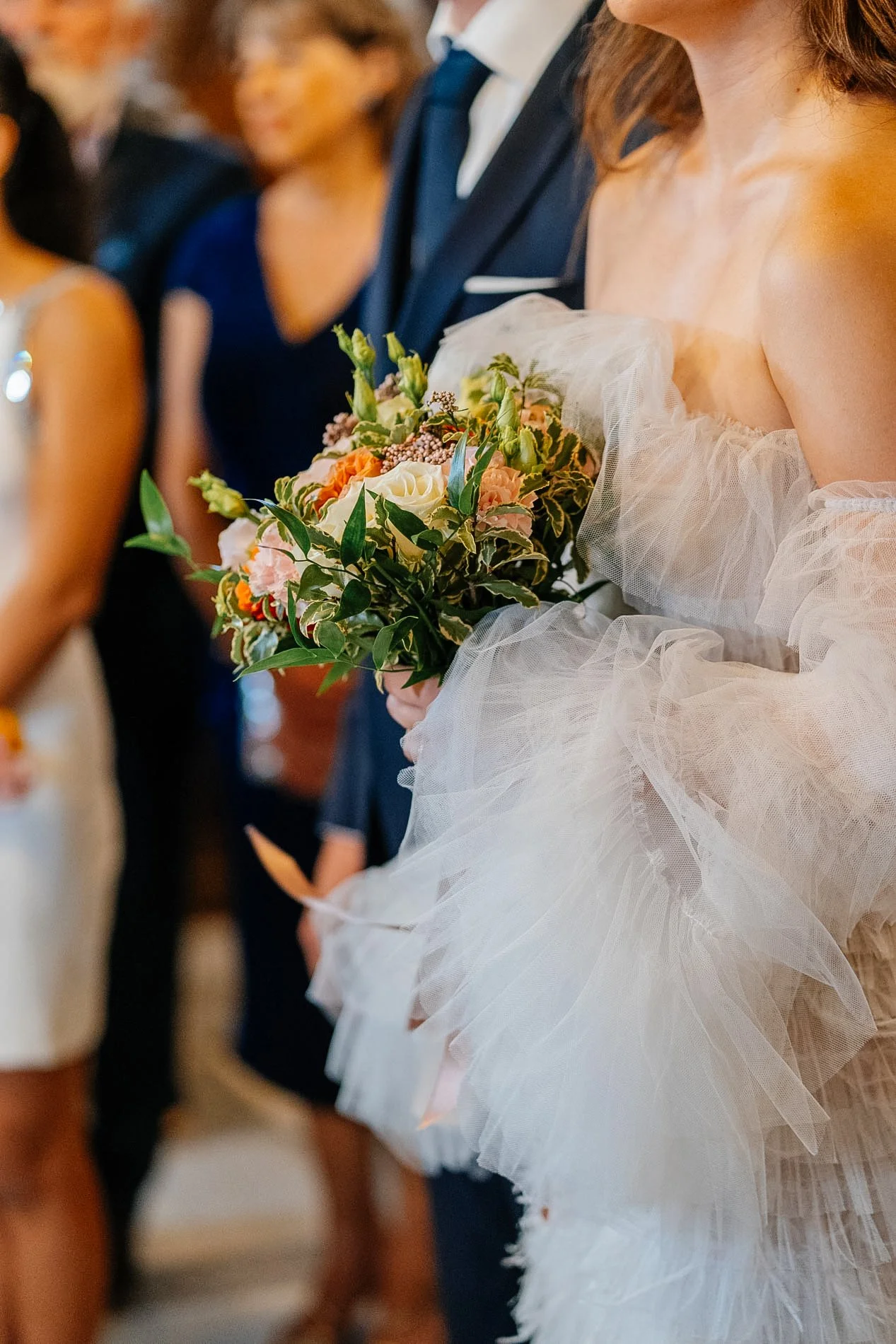 A bride holding a bouquet of flowers during a wedding ceremony, with guests blurred in the background.