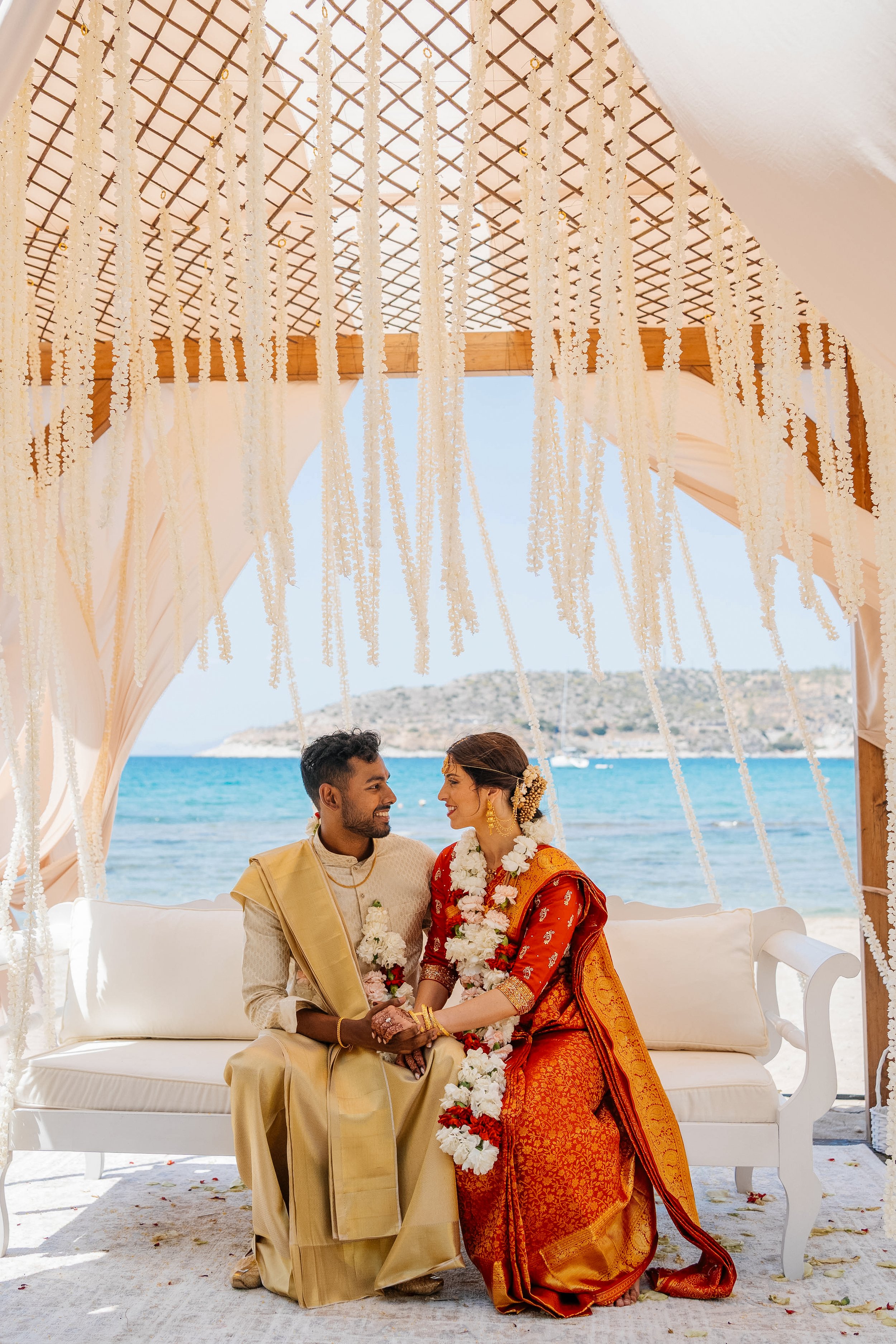 A couple in traditional Indian attire exchanging wedding vows at a beachside wedding ceremony under a decorated canopy with a view of the ocean and distant hills.