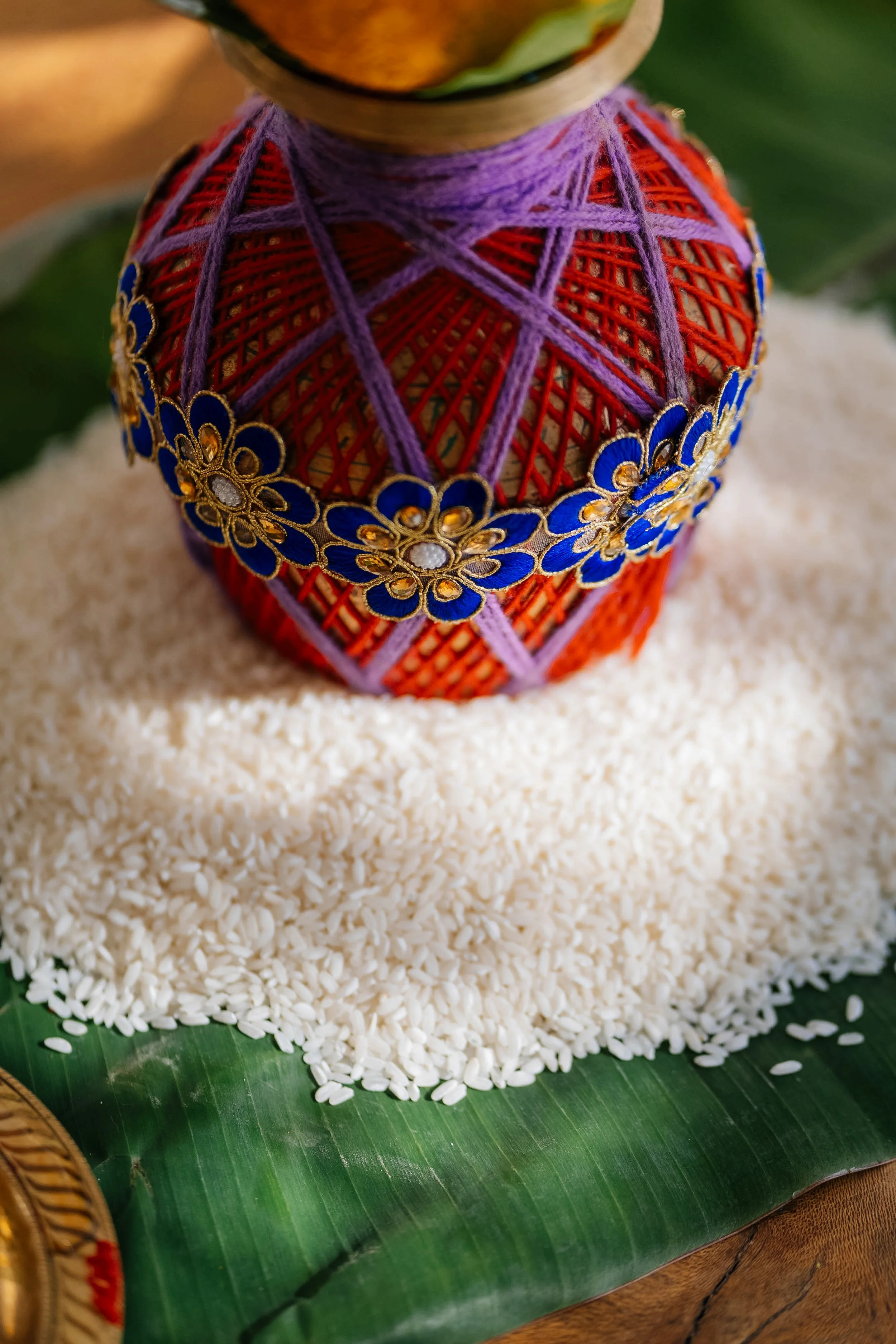A traditional Thai decorative container made of woven materials with colorful embellishments, placed on a large green leaf, surrounded by rice grains.