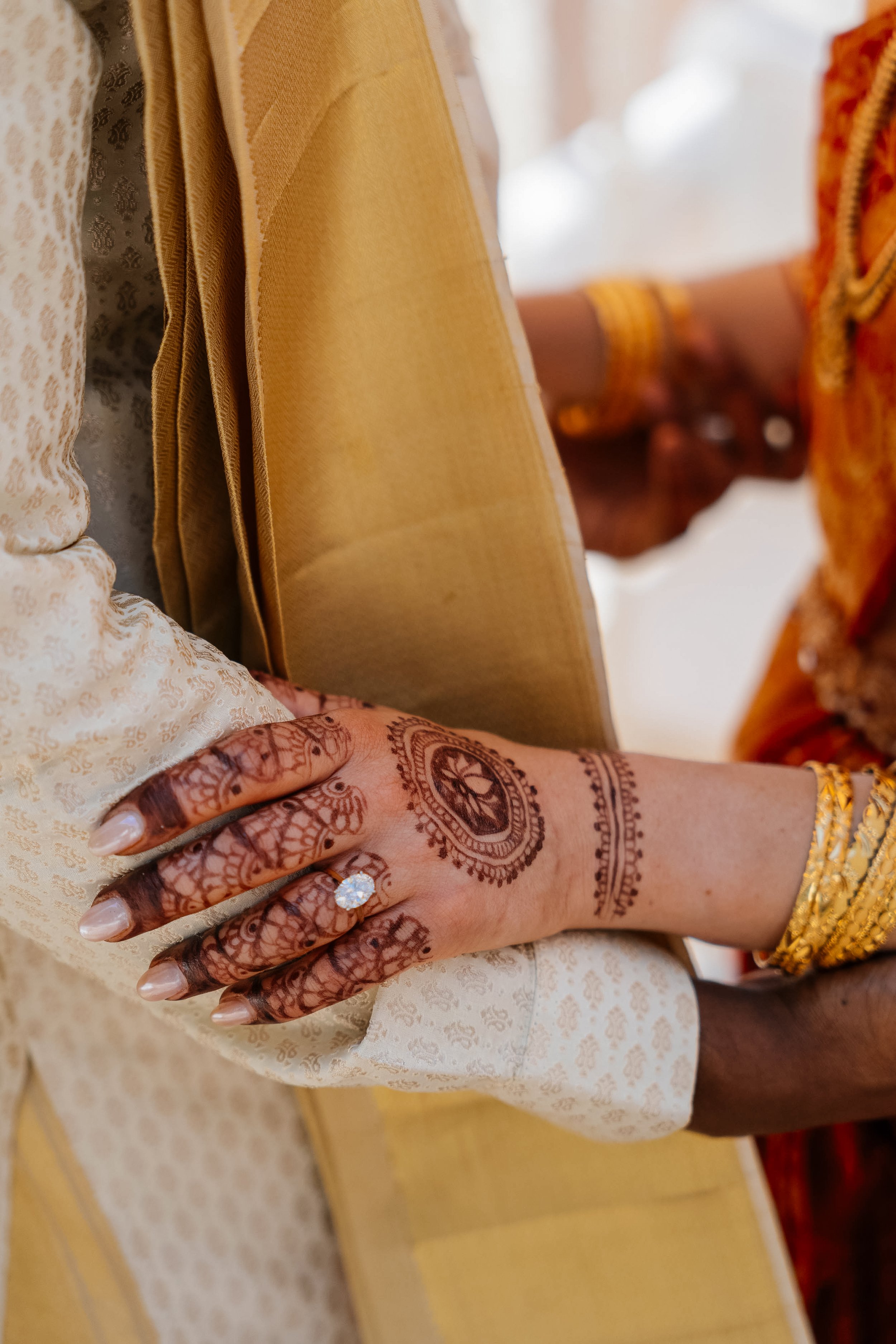 Close-up of a woman's hand with henna designs, wearing gold rings and bracelets, resting on cream-colored fabric, with traditional clothing in the background.