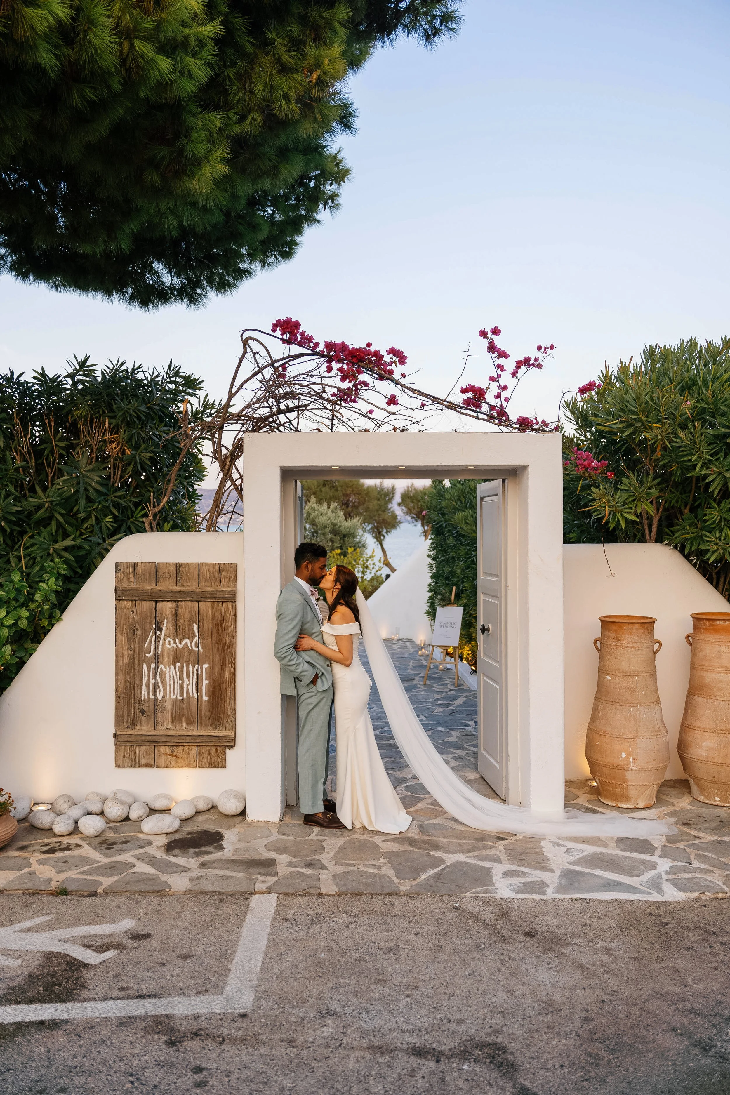 A newlywed couple stands close together, sharing a kiss under a white archway in an outdoor courtyard with a sign that reads 'Hand Residence'. The bride is wearing a white wedding dress with a long train, and the groom is in a light gray suit. The ba