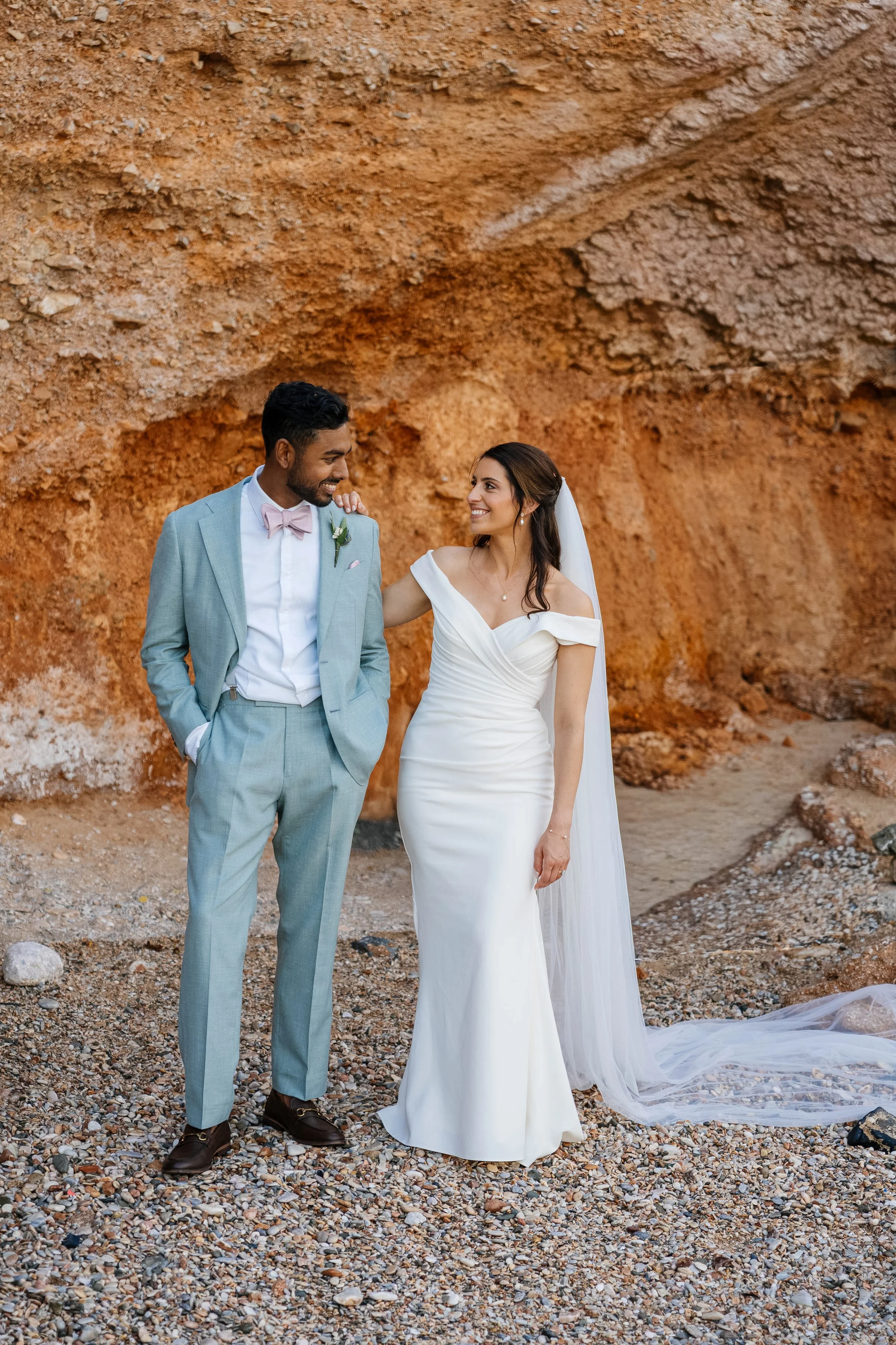 A newlywed couple dressed in wedding attire, standing on a rocky terrain with a large orange rock formation in the background, smiling and looking at each other.