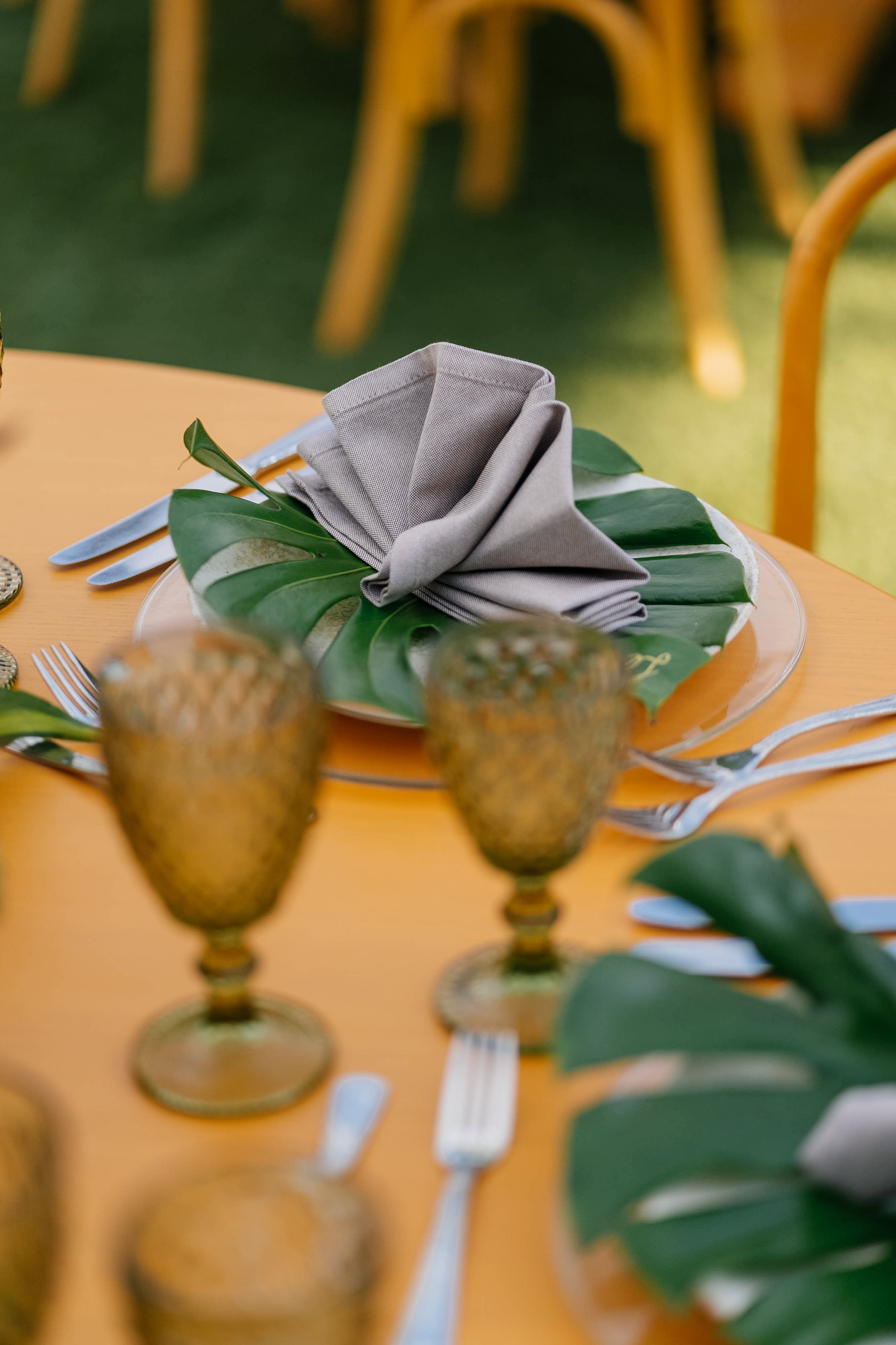 Table setting with a folded gray napkin, green leaves, and glass goblets on a yellow table.