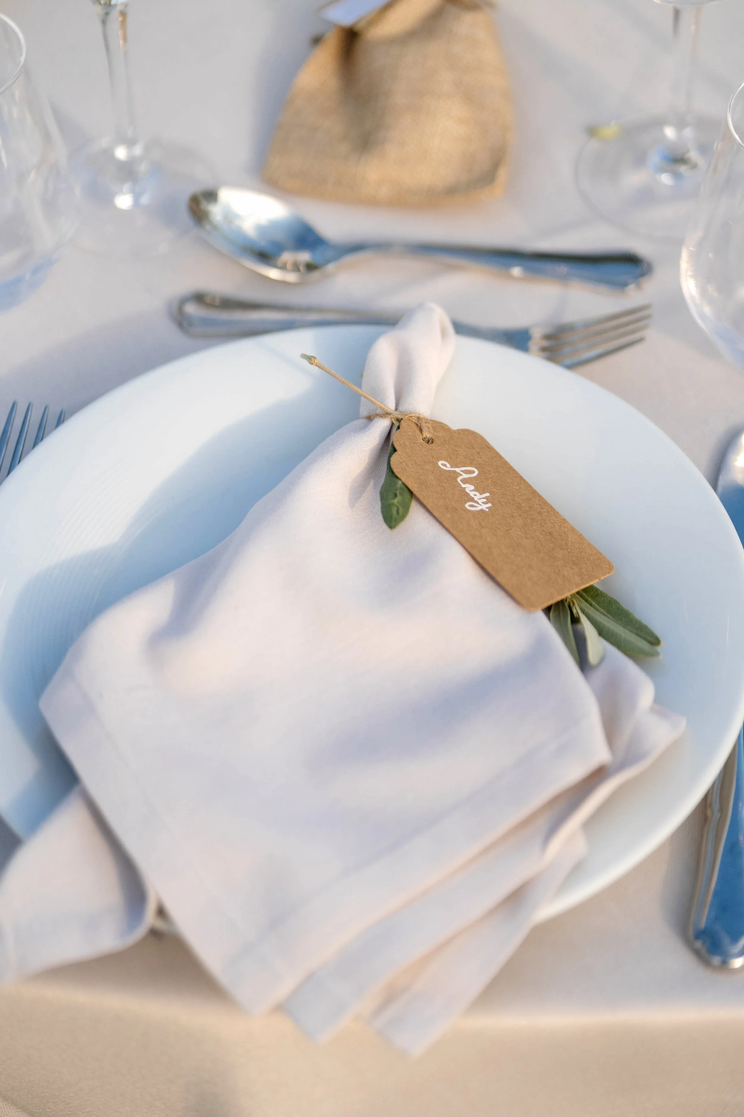 A white napkin wrapped around cutlery with a small brown paper tag and green leaves, placed on a white plate at a formal dining setting.