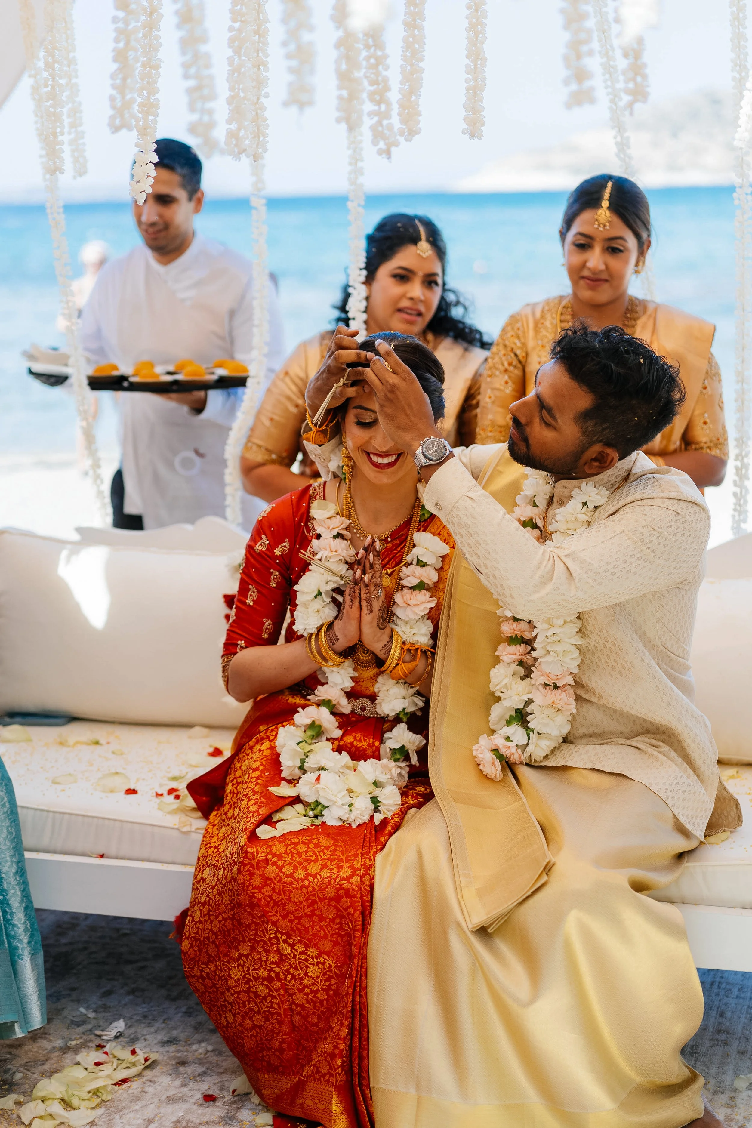 A traditional Indian wedding ceremony featuring a bride and groom seated on the ground, exchanging garlands, with women and a man in traditional attire around them. The background shows the ocean, indicating a beach wedding.