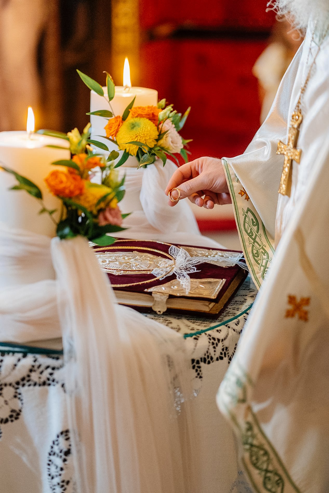 Close-up of a person dressed in religious attire placing a ring on someone's finger during a wedding ceremony, with candles, flowers, and a prayer book on a decorated table.