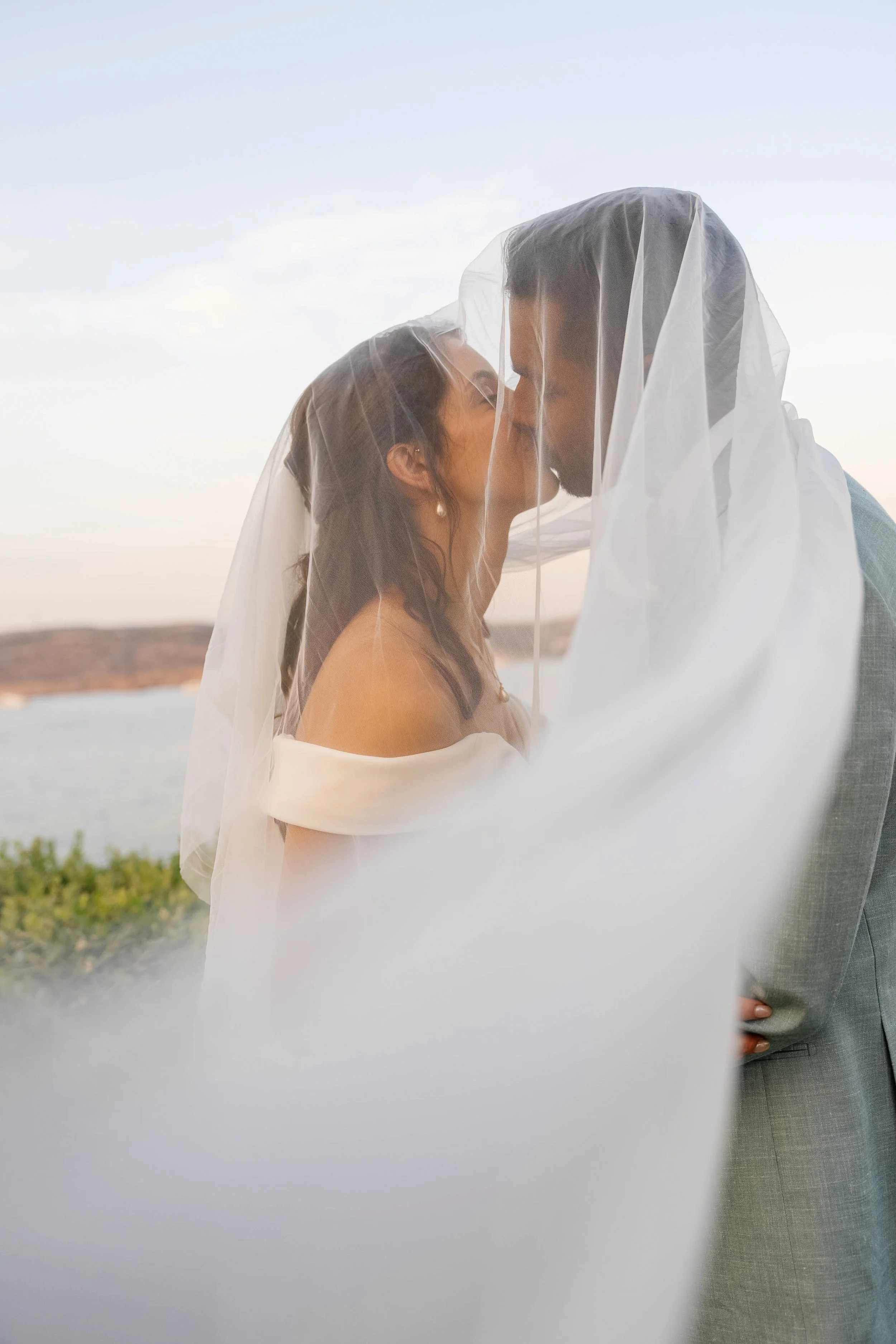 A couple sharing a kiss, partially covered by a sheer fabric, outdoors during sunset or sunrise, with a landscape and water in the background.