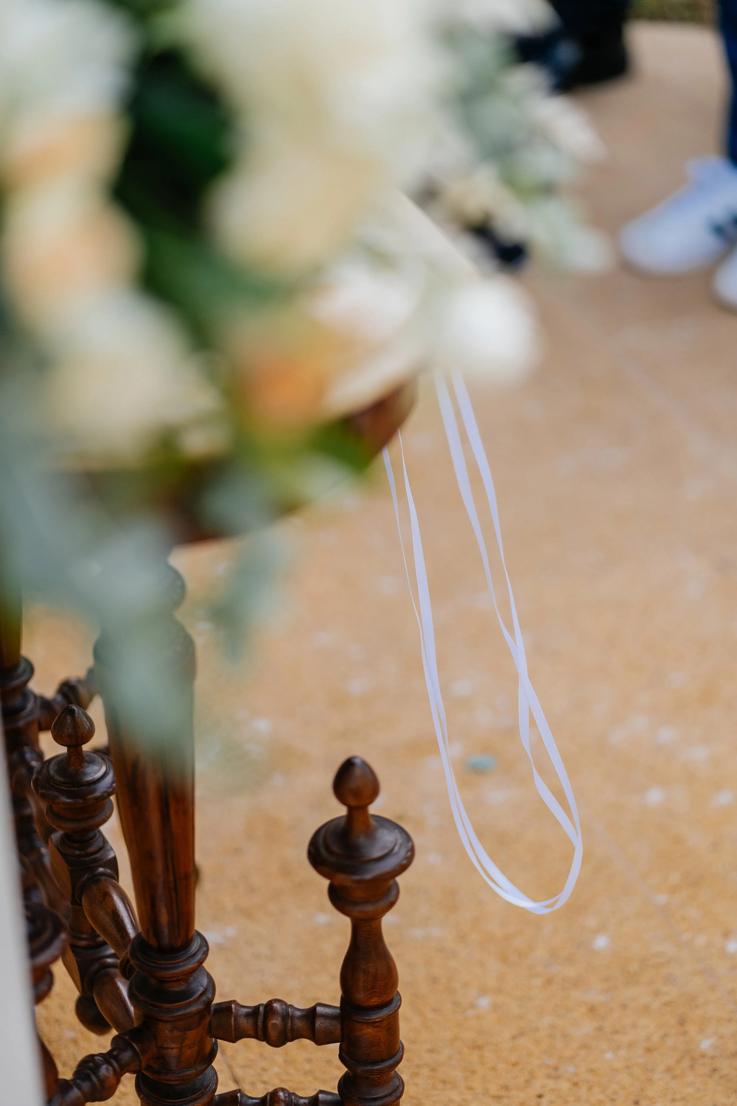 Close-up of a row of small, dark wooden spindles on a piece of furniture, with a blurred background featuring light-colored flooring and some shoes, and a partially blurred object with thin white cords hanging.