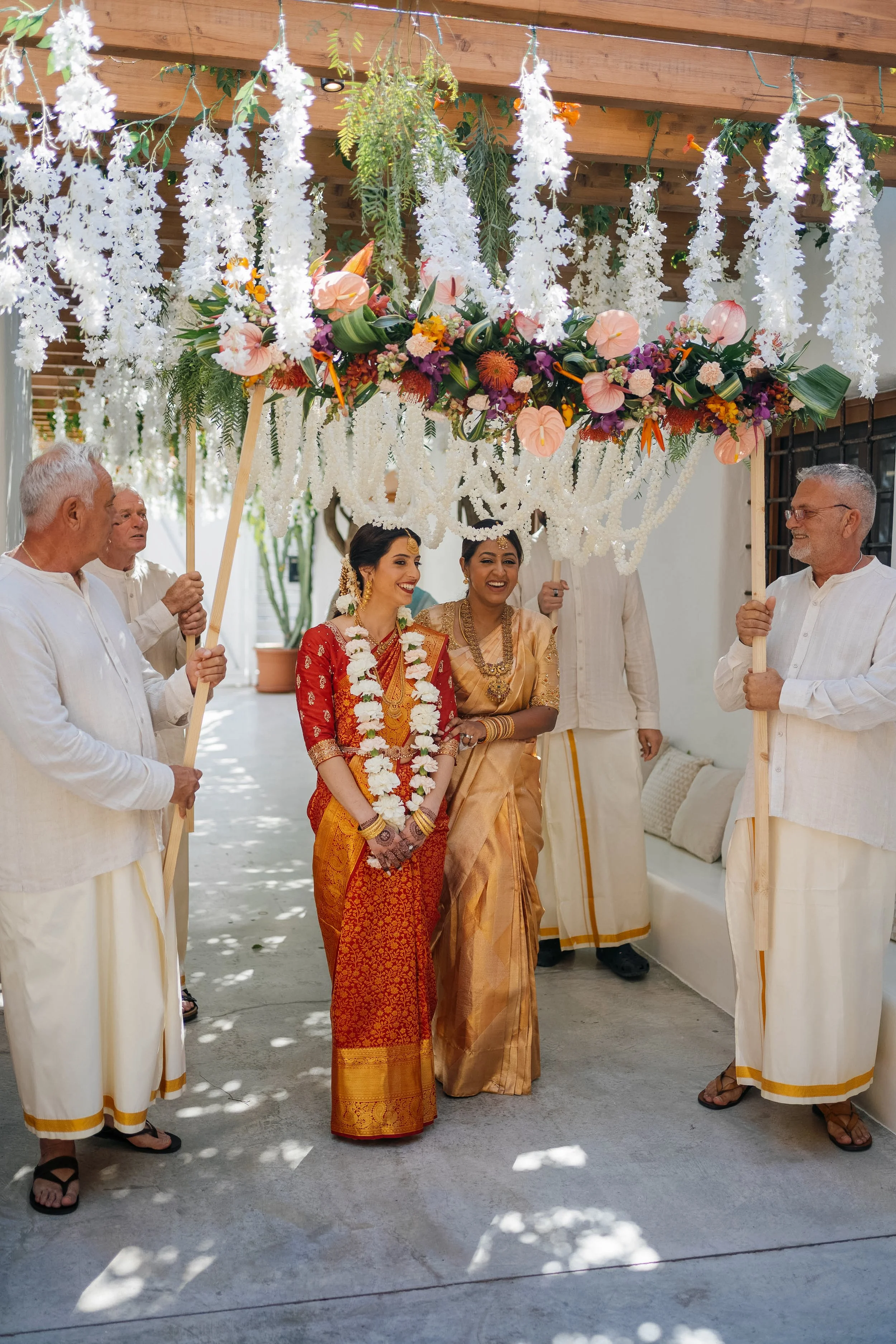 A group of four men in white traditional Indian clothing holding a floral canopy over two women dressed in traditional Indian attire, during a celebration or wedding ceremony outdoors.