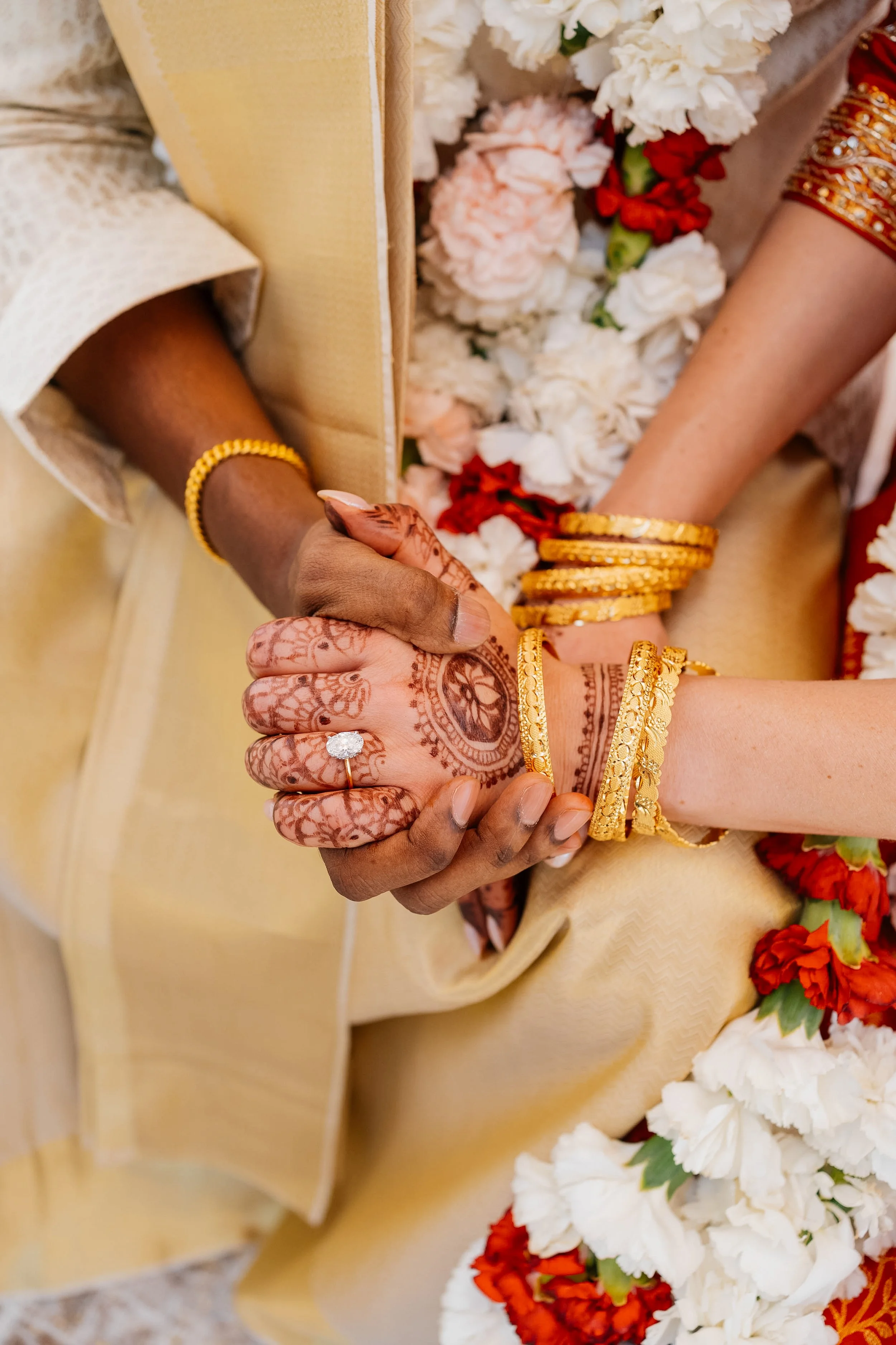 Close-up of two hands holding each other, adorned with traditional gold jewelry, during a wedding ceremony with white and red flower decorations in the background.