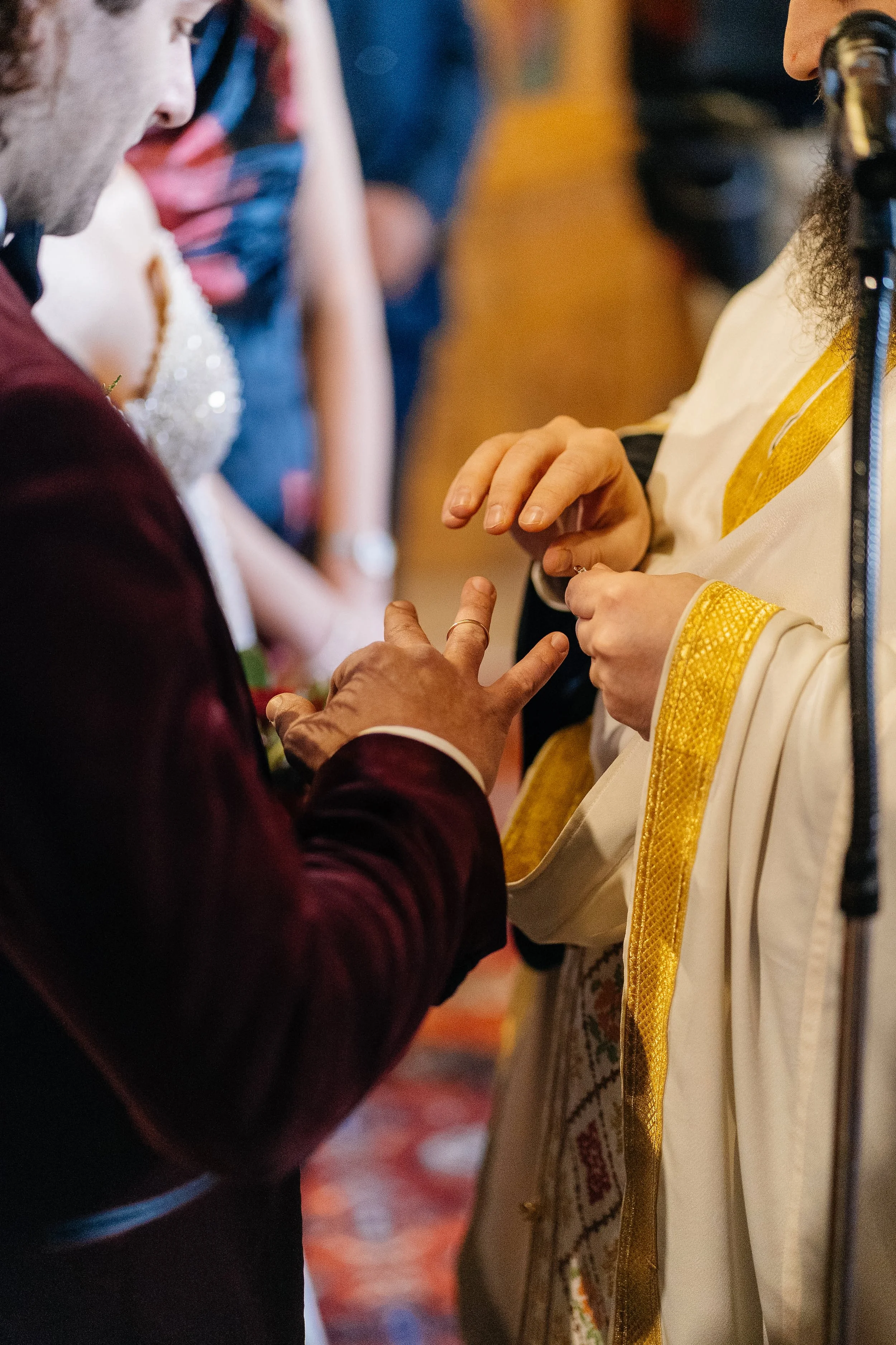 Close-up of a couple exchanging wedding rings during a wedding ceremony. The bride is wearing a white dress with gold accents, and the groom is dressed in a dark suit.