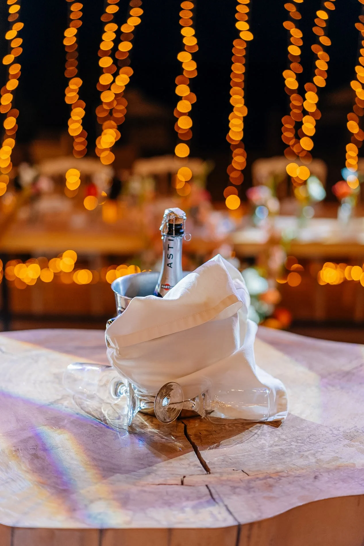 A bottle of sparkling wine in an ice bucket on a table with a white cloth, in front of a background with yellow string lights.