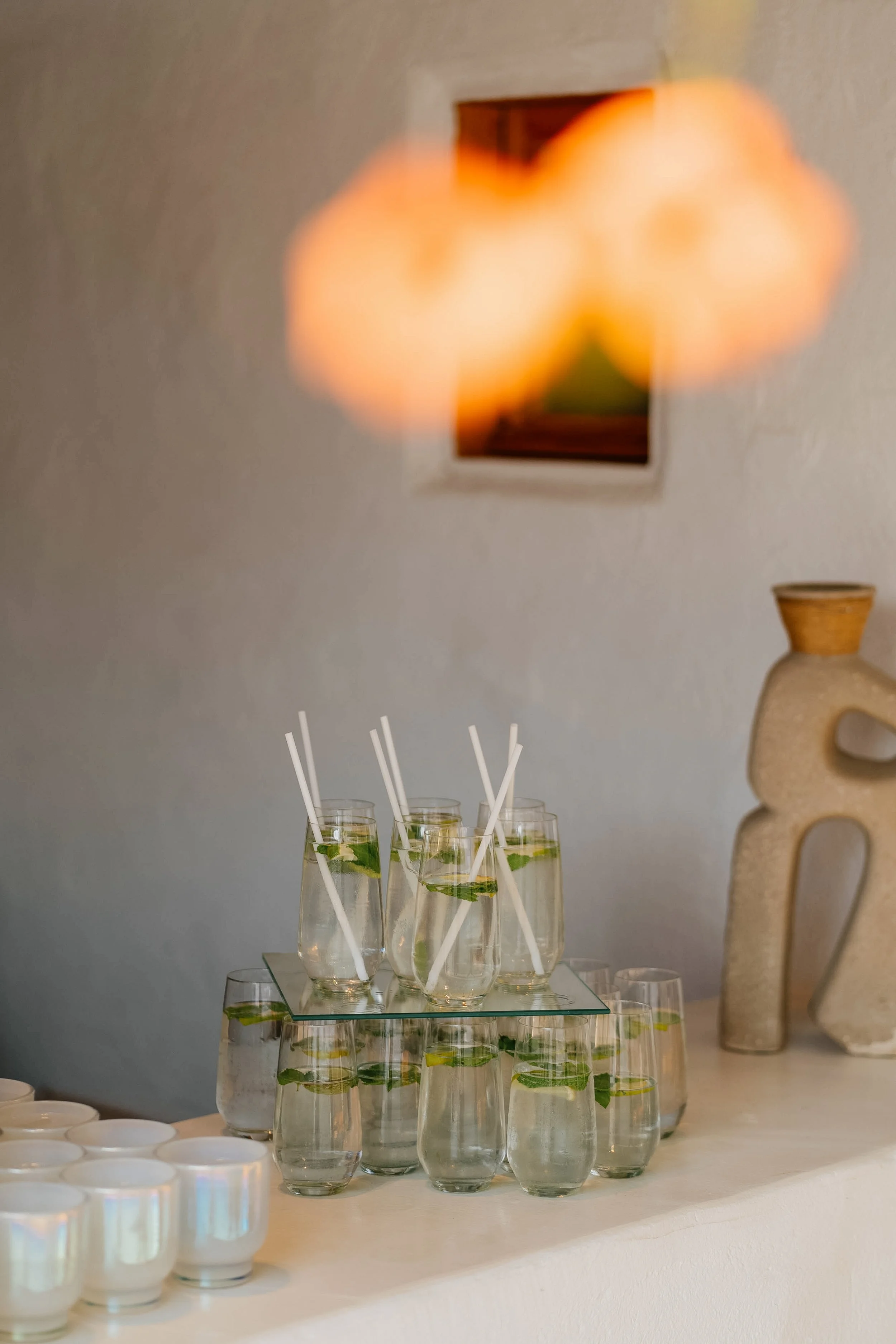 A display of multiple glasses of lemonade with mint leaves and straws on a glass tray, placed on a table. In the background, a large decorative ceramic vase and a wall art piece are visible.