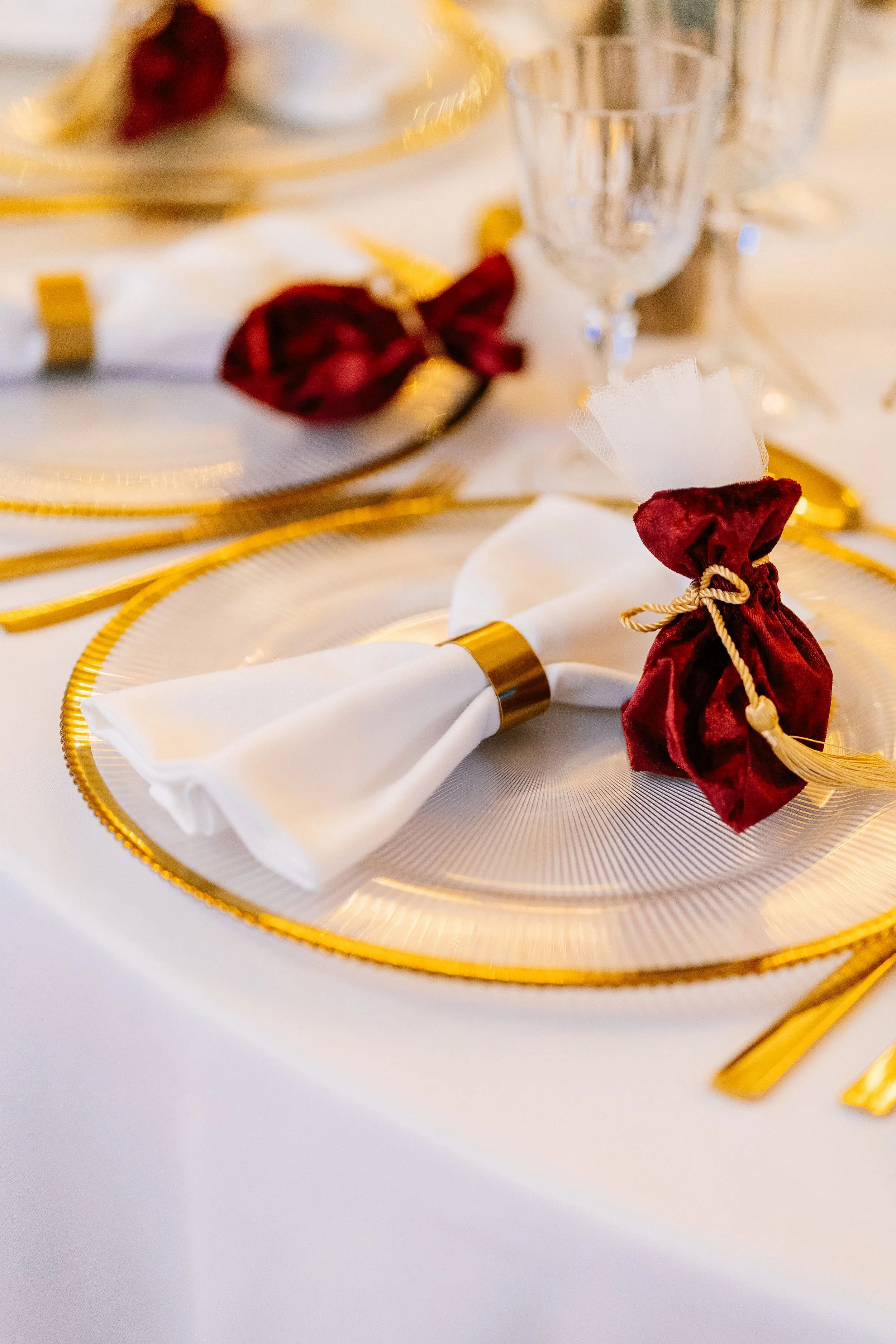 Elegant table setting with white napkin wrapped in gold and red velvet ties, gold-rimmed plates, a clear glass, and a festive decor theme with gold accents.
