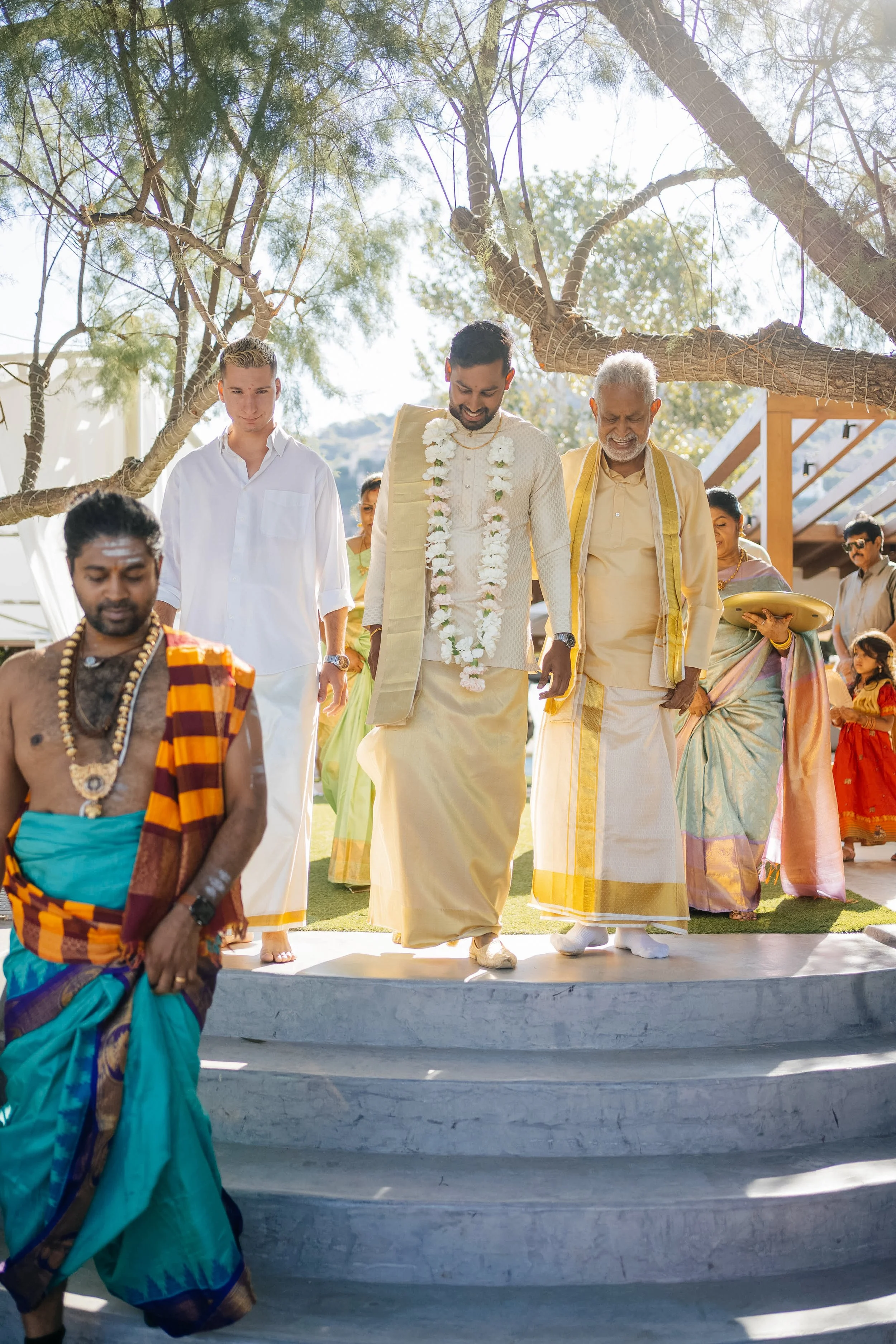 Indian groom and elder are ascending steps during a traditional wedding ceremony, surrounded by family members under a tree.