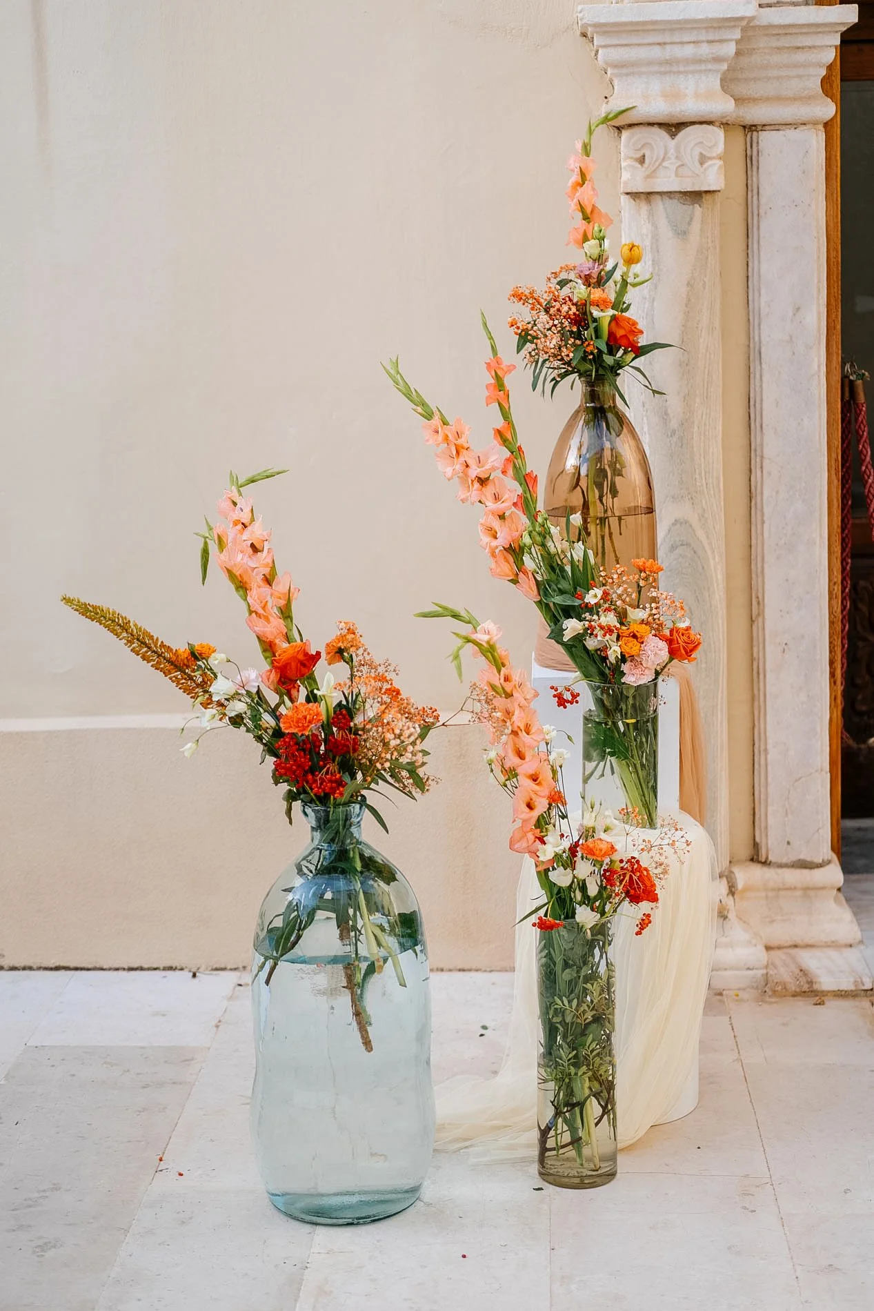 Three vases with colorful flowers placed on a white cloth on the ground against a beige wall and stone column.