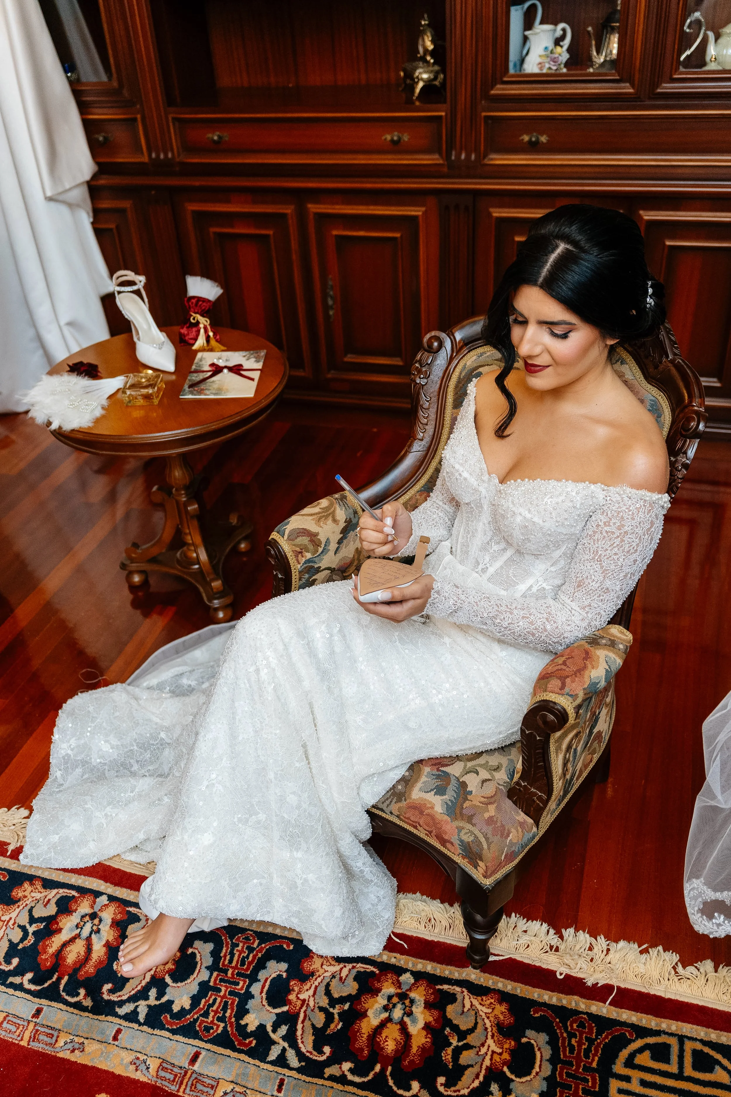 A woman in a white wedding gown sitting on an ornate wooden chair, signing a document, in a room with wooden cabinets and a patterned rug.