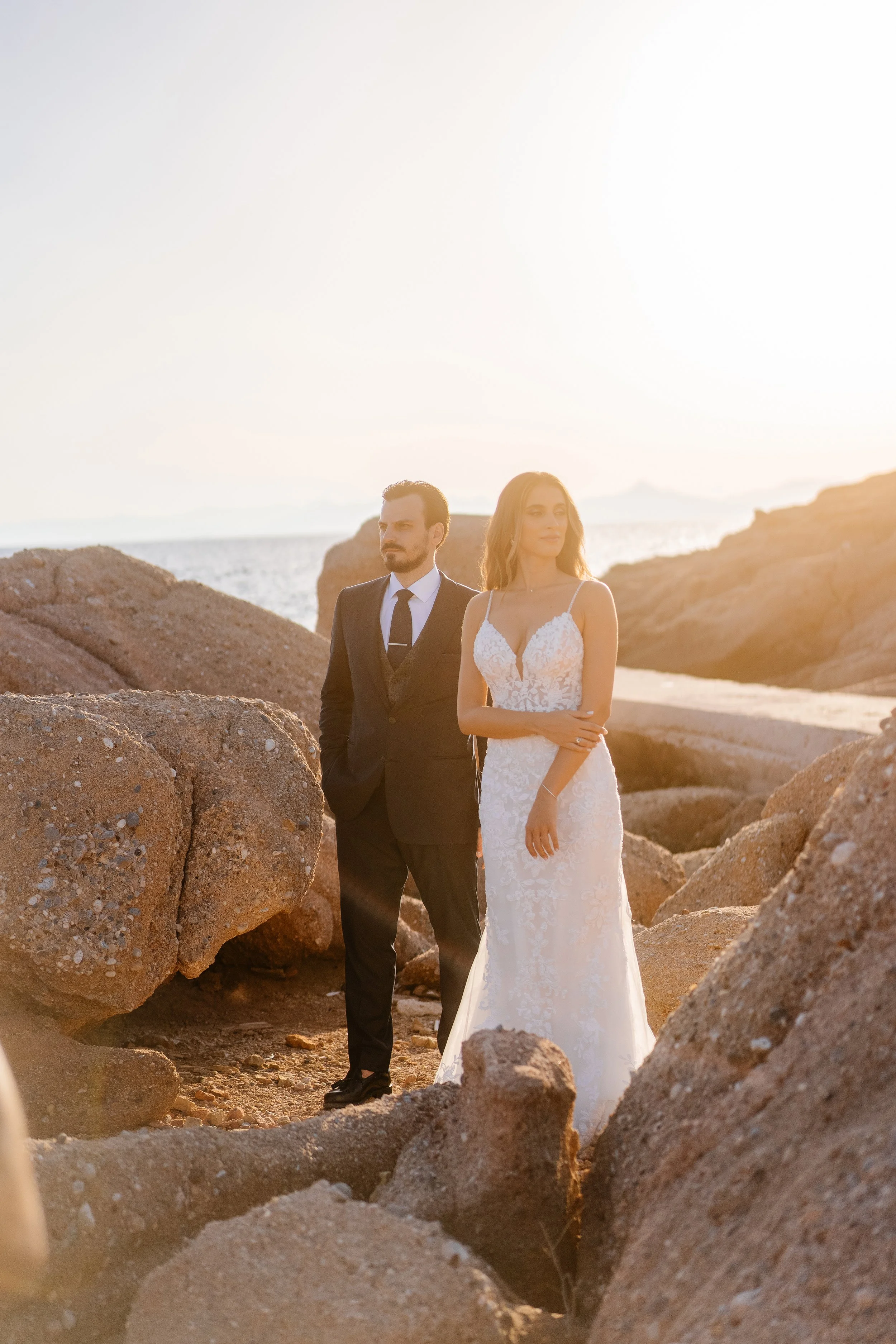 A man in a black suit and tie and a woman in a wedding dress standing among large rocks on a beach during sunset.