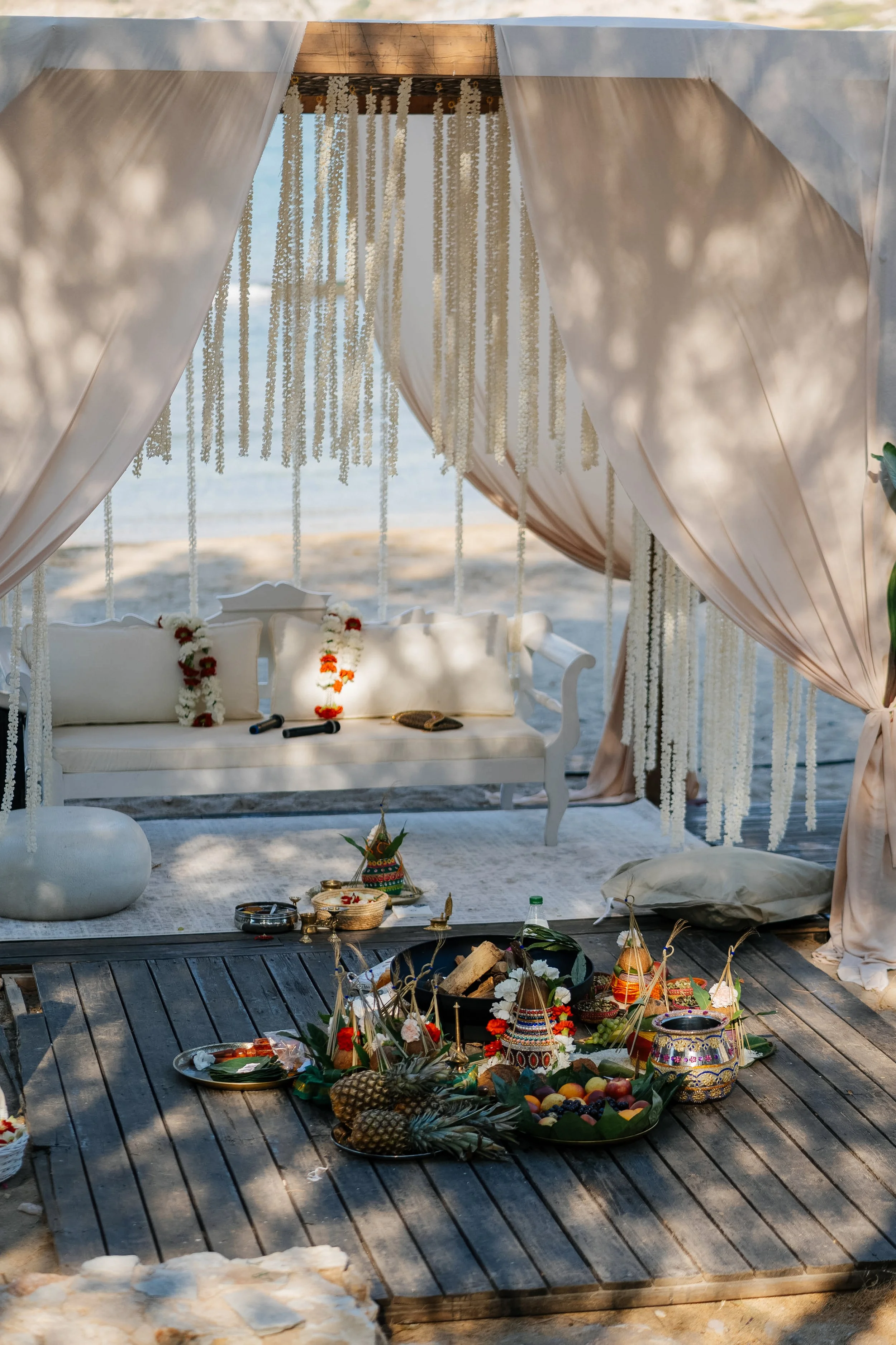 A traditional Hindu or Indian wedding setup on a beach, with a white decorated sofa, floral garlands, and colorful offerings placed on a wooden platform.