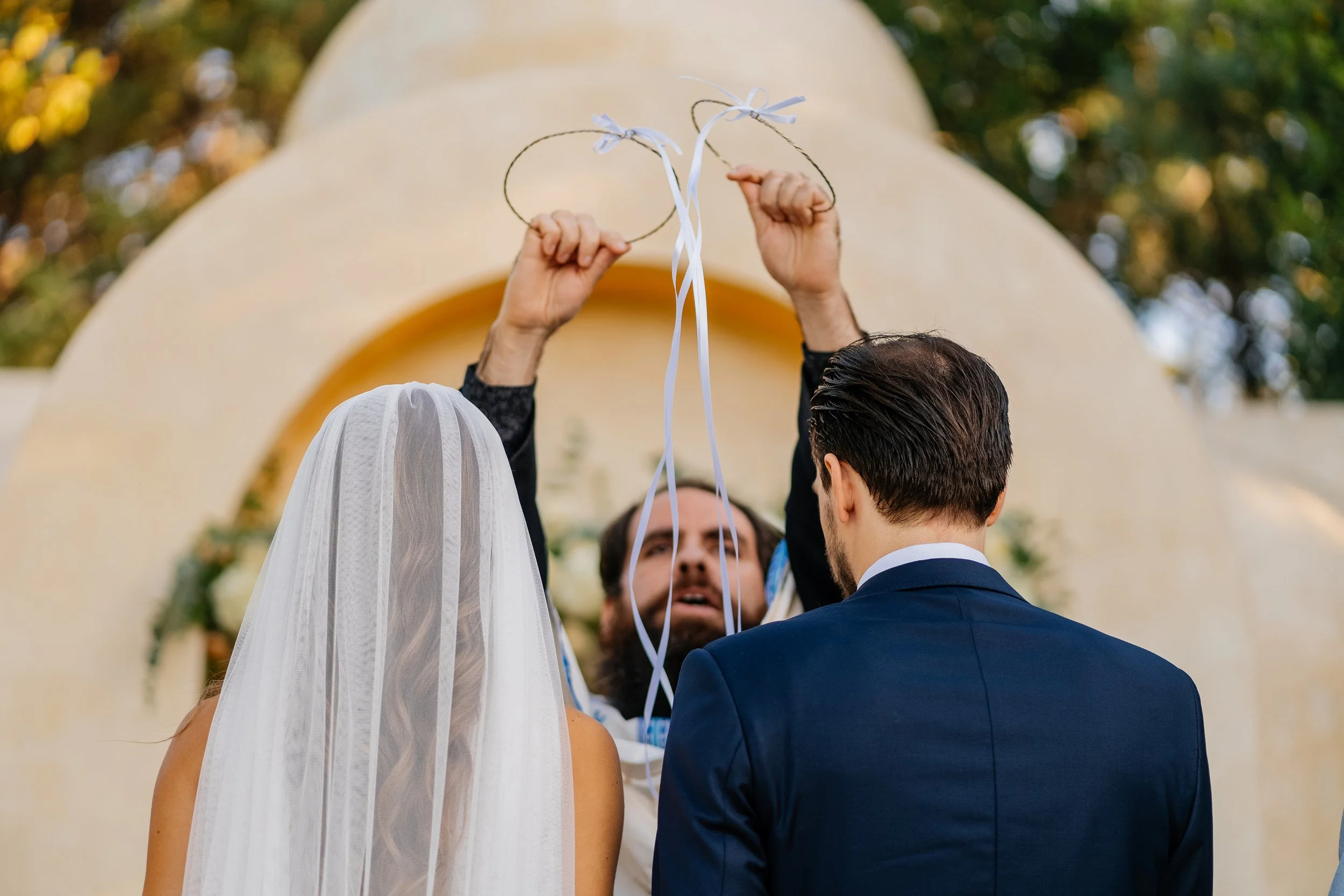 A couple getting married outdoors, raising two wedding rings on a white ribbon tied to sticks, with a blurred arch and greenery in the background.