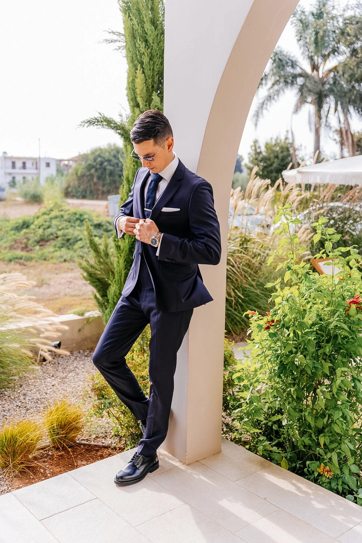 A young man in a dark suit, white shirt, and tie standing outdoors near a beige architectural column. He is wearing sunglasses, a wristwatch, and black dress shoes, and is adjusting his suit jacket. He is posing with one foot slightly forward and his