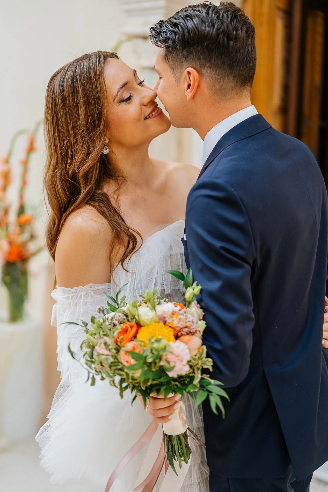 A bride and groom are close, about to kiss, during their wedding. The bride has long, wavy brown hair and is holding a colorful bouquet. The groom has short, styled dark hair and is wearing a navy blue suit. They are indoors with blurred background d