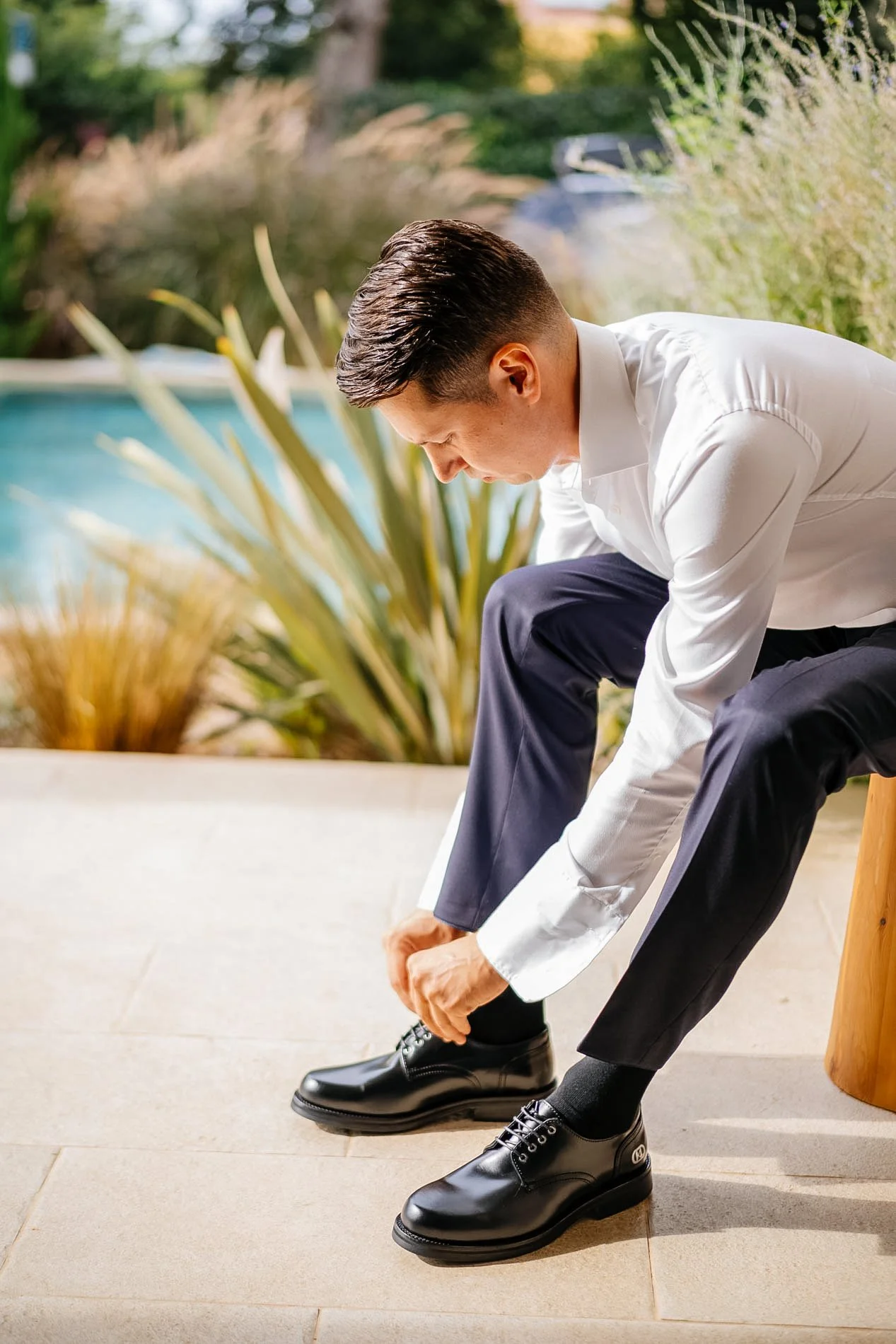 A man dressed in a white shirt and dark pants tying his black dress shoes outdoors during daytime.