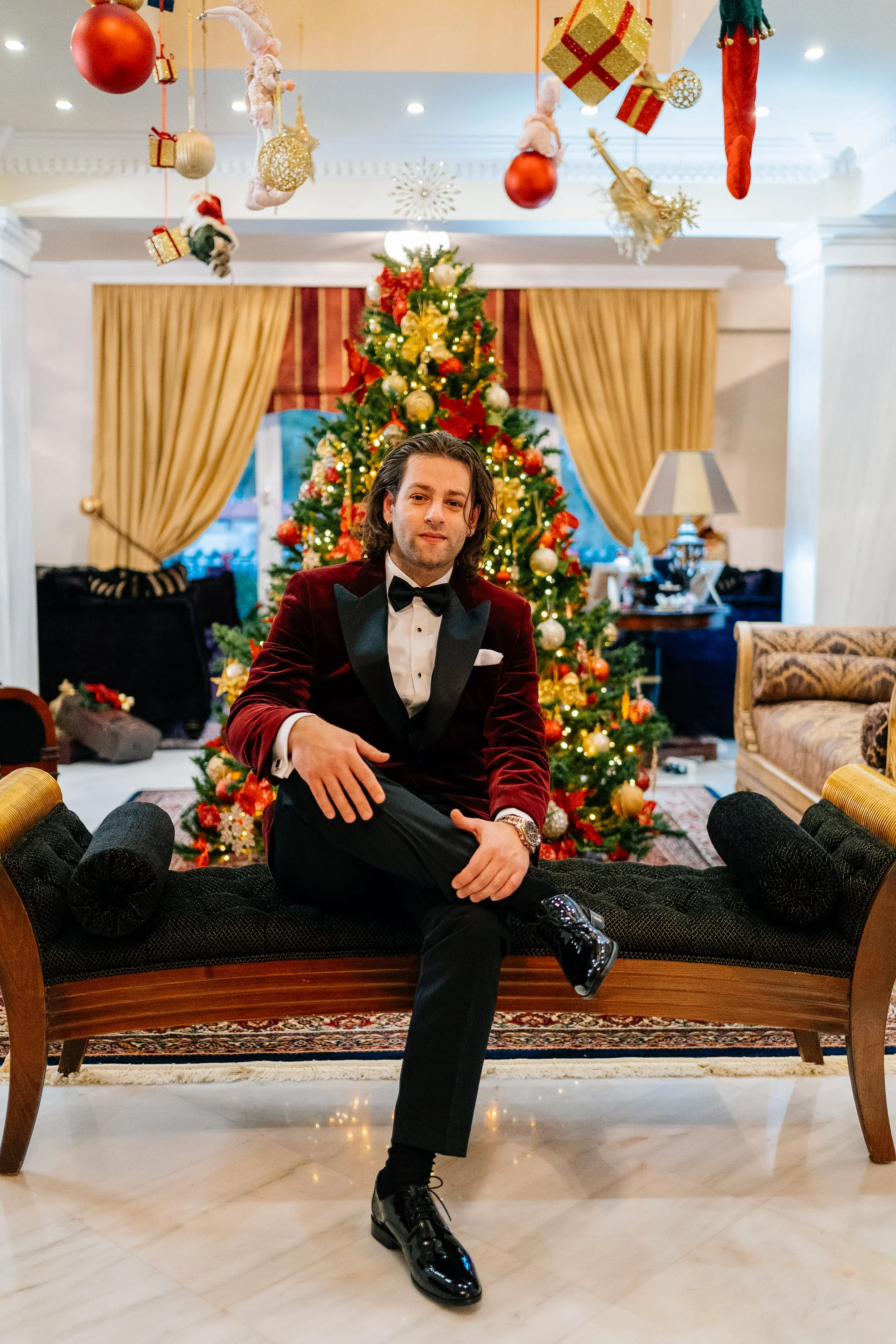 A man in a tuxedo sitting on an elegant black and wooden couch in a decorated room with a Christmas tree, gold and red ornaments, and hanging holiday decorations including Christmas stockings and wrapped presents.