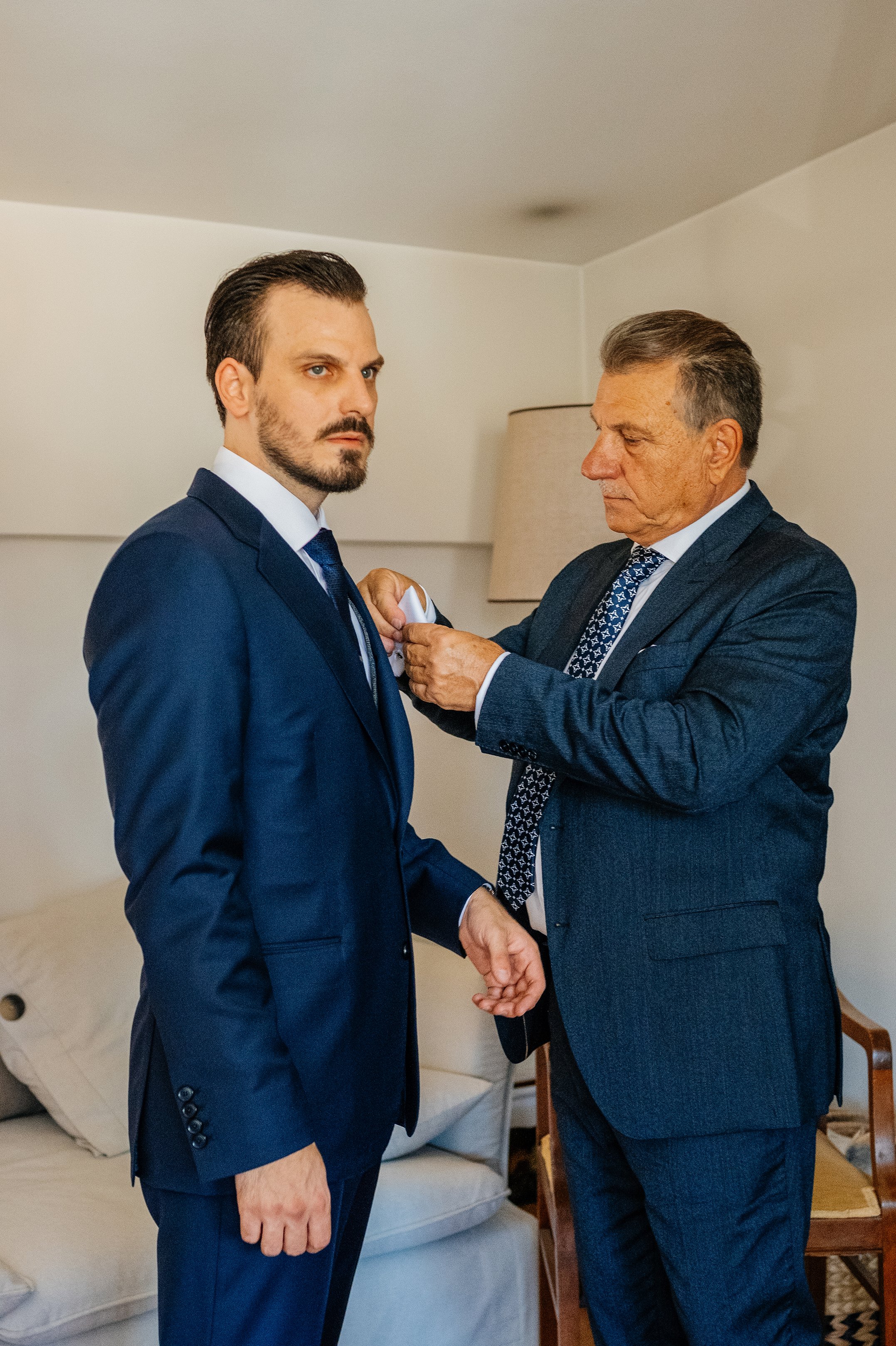 Two men in suits are in a room, with one man helping the other adjust his cufflinks. One man has dark hair and a beard, the other has gray hair. They are standing near a white sofa and a lamp.