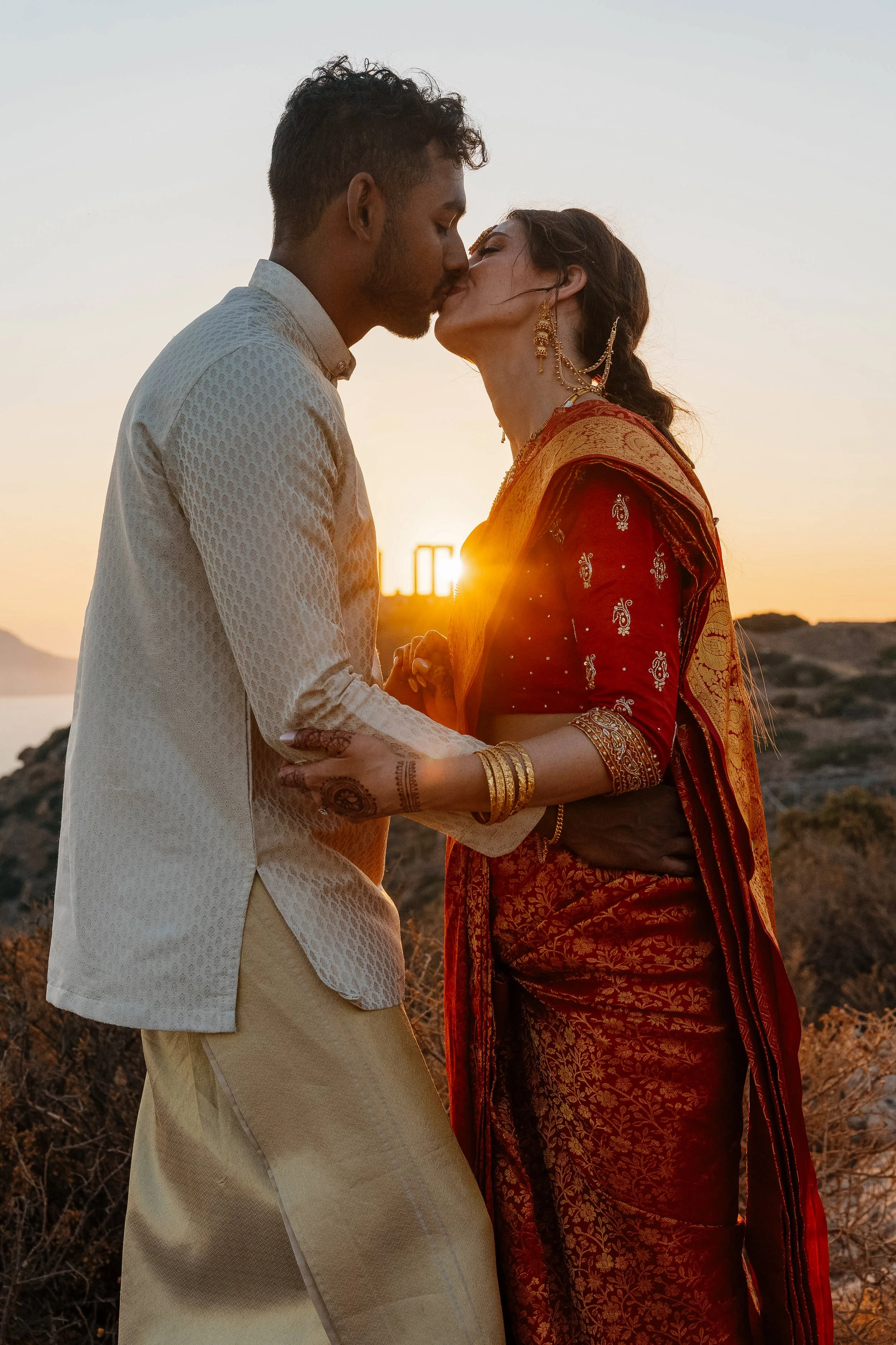 A couple kissing at sunset on a rocky outdoor hillside, with the sun setting behind them.