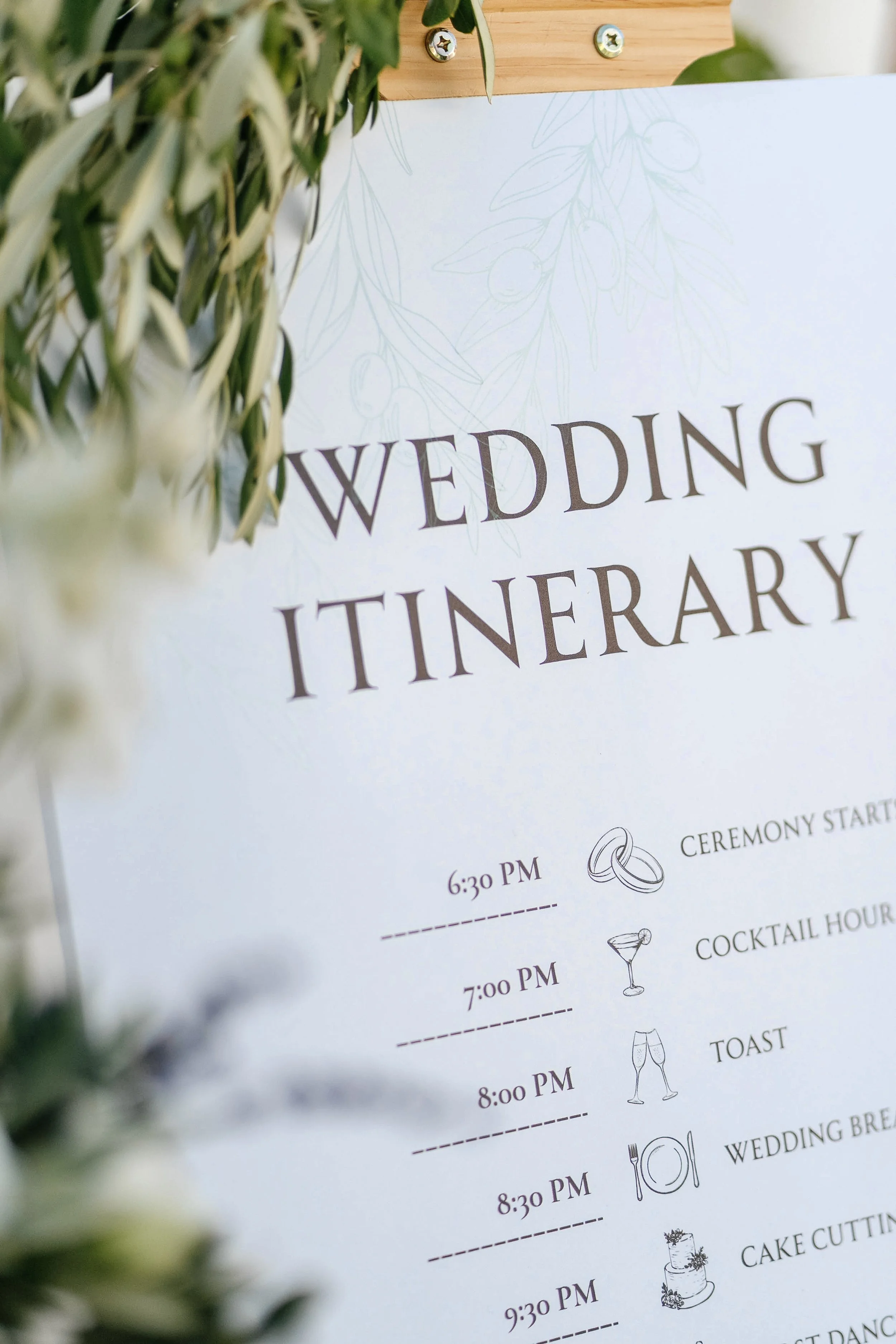 A wedding itinerary with a white background and black text, decorated with light illustrations of leaves and olives, partially obscured by green foliage in the foreground.
