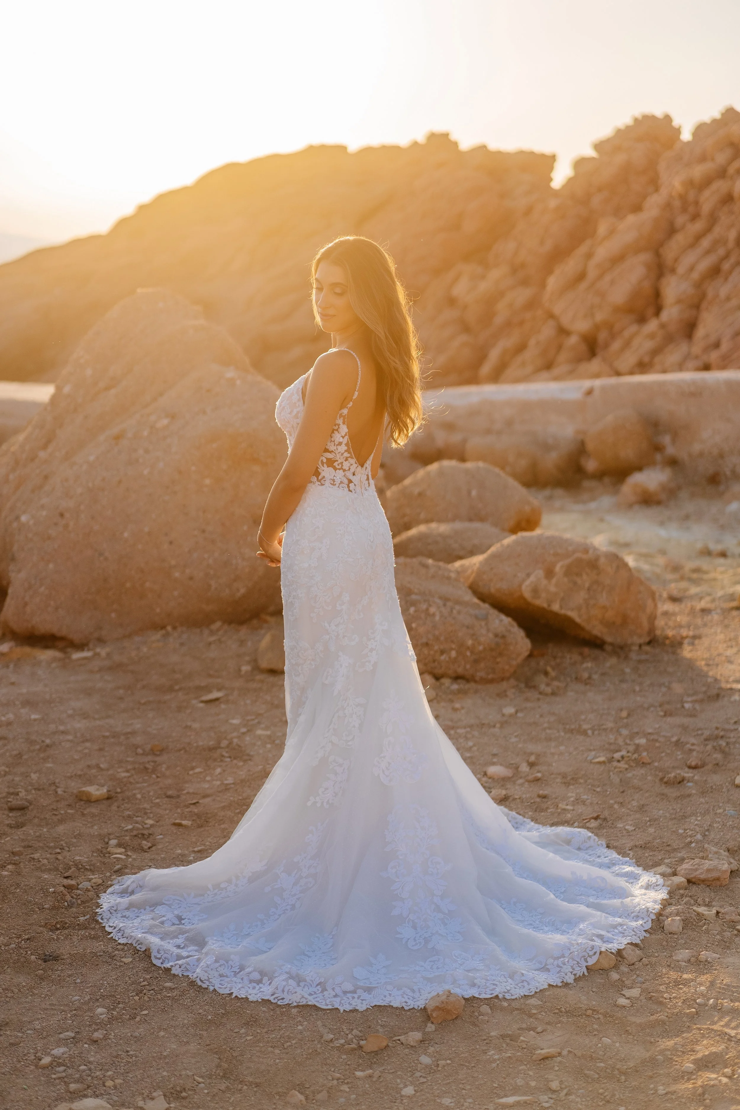 A woman in a white lace wedding dress standing outdoors in a desert area with rocky hills behind her, during sunset.