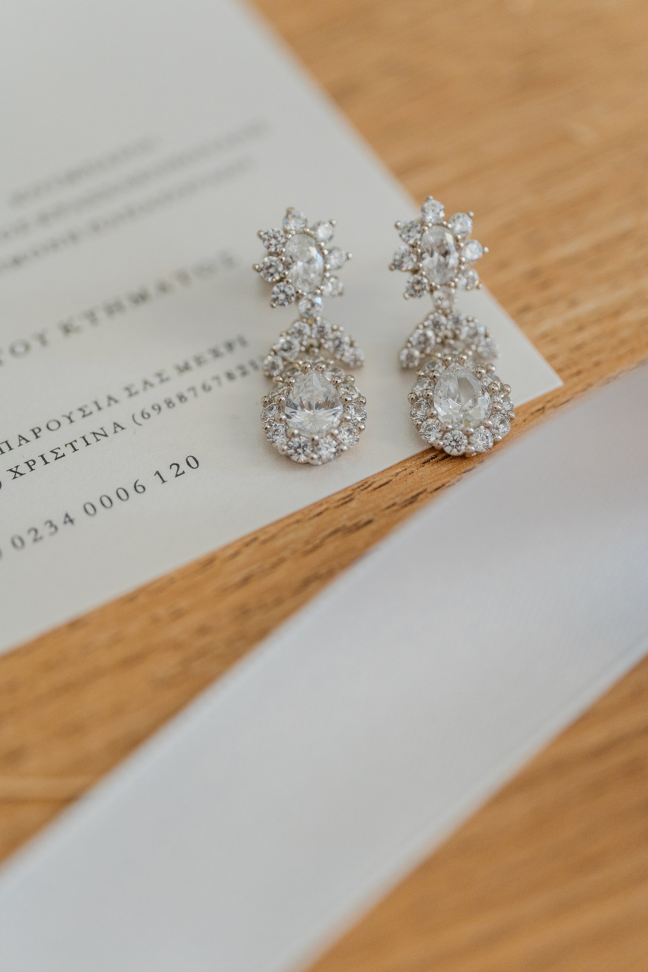 Pair of silver earrings with large central clear gemstones surrounded by smaller gemstones, placed on a white paper with printed text, on a wooden surface.