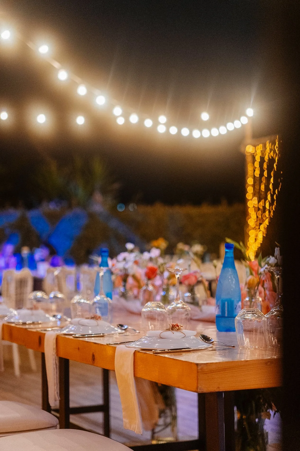 Decorated outdoor dining setup with a long table covered with a pink tablecloth, floral arrangements, and tableware, under string lights at night.